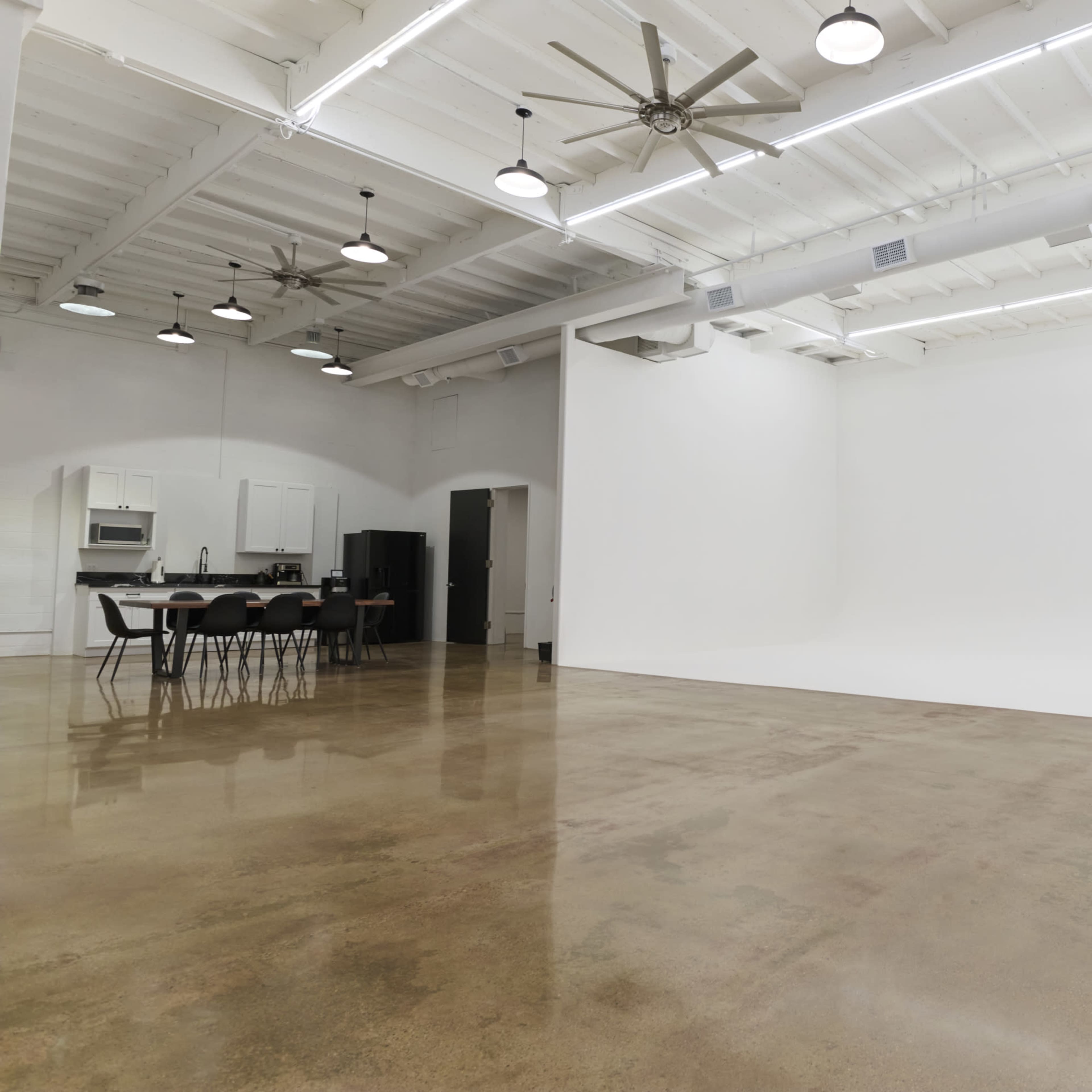 A spacious, well-lit studio with a clean white backdrop, polished concrete floor, and a kitchen area with a dining table and chairs in the corner.