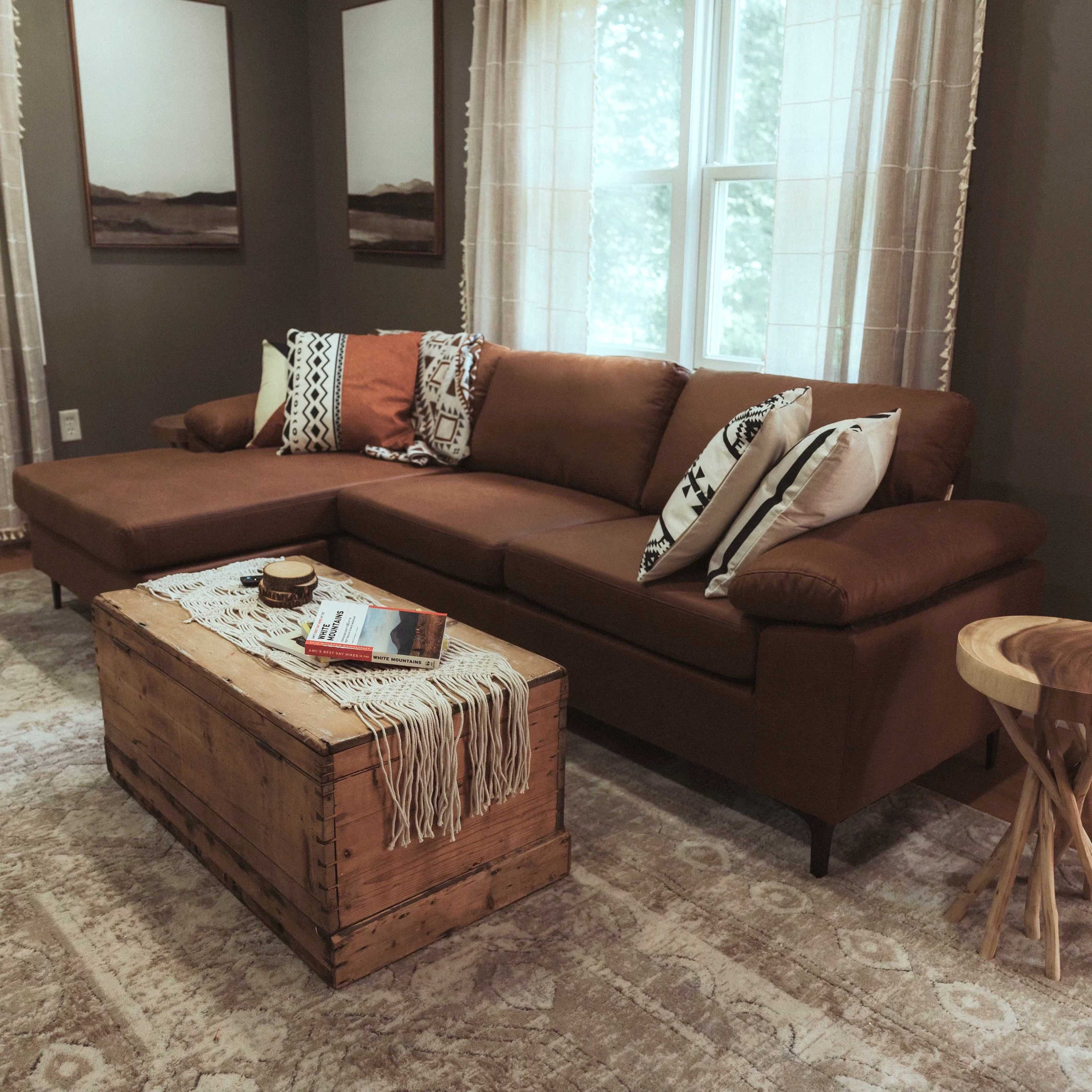 A brown sectional sofa with patterned pillows and a wooden coffee table in a cozy living room with large windows dressed in sheer curtains.