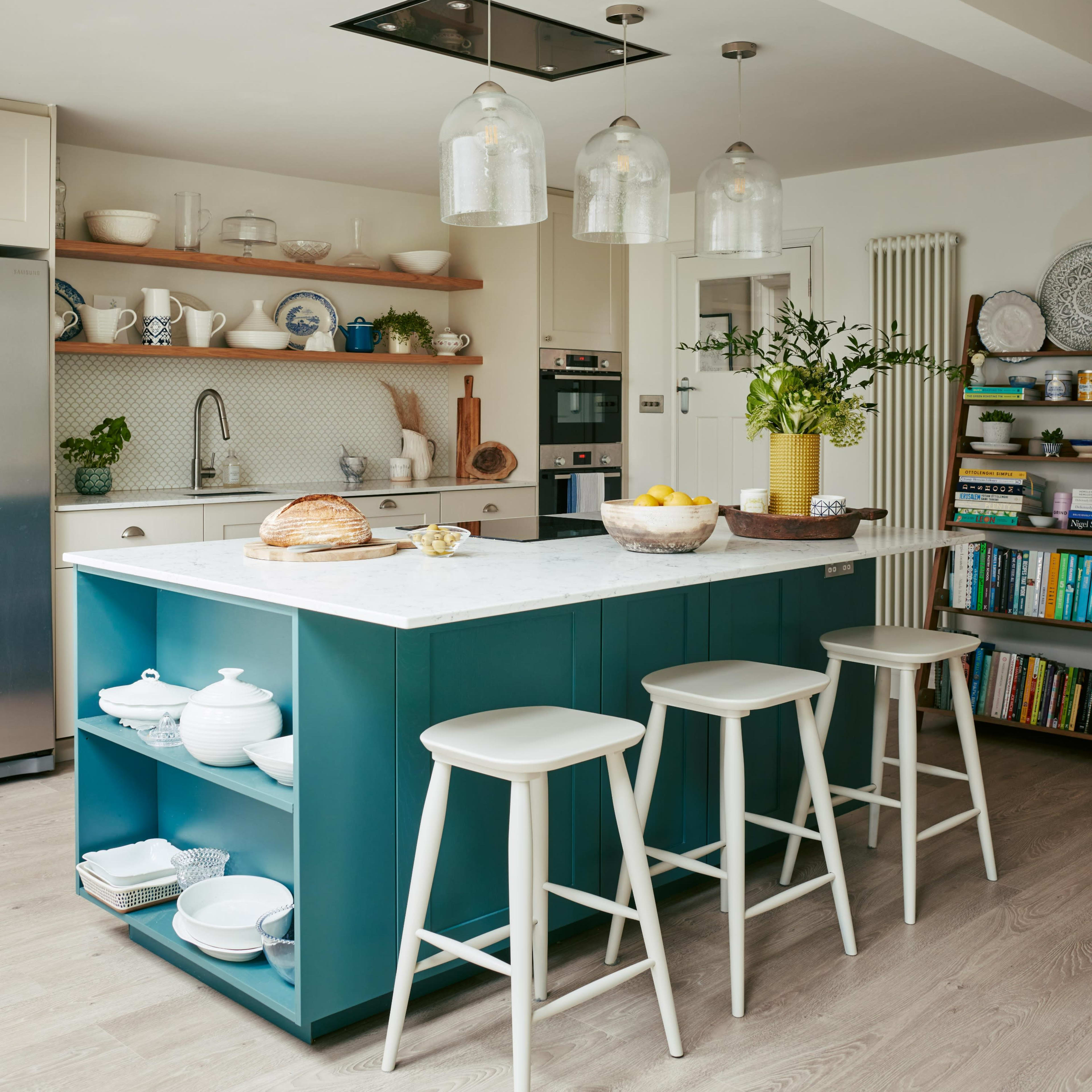 A modern kitchen features a blue island with a marble countertop, three white stools, and open shelving displaying dishes and decor.