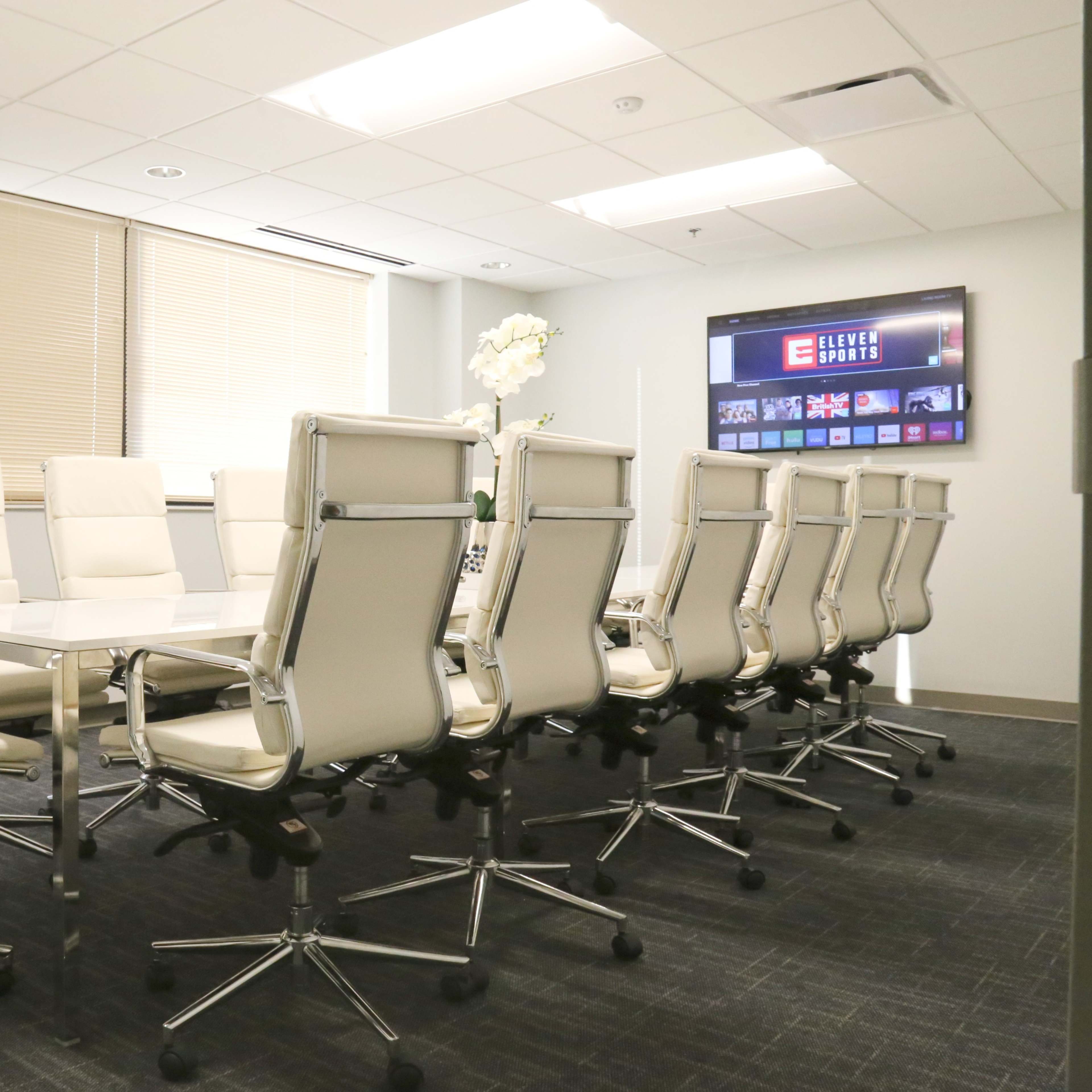 A conference room features a long glass table surrounded by twelve white ergonomic chairs and a large screen on the wall displaying media content.