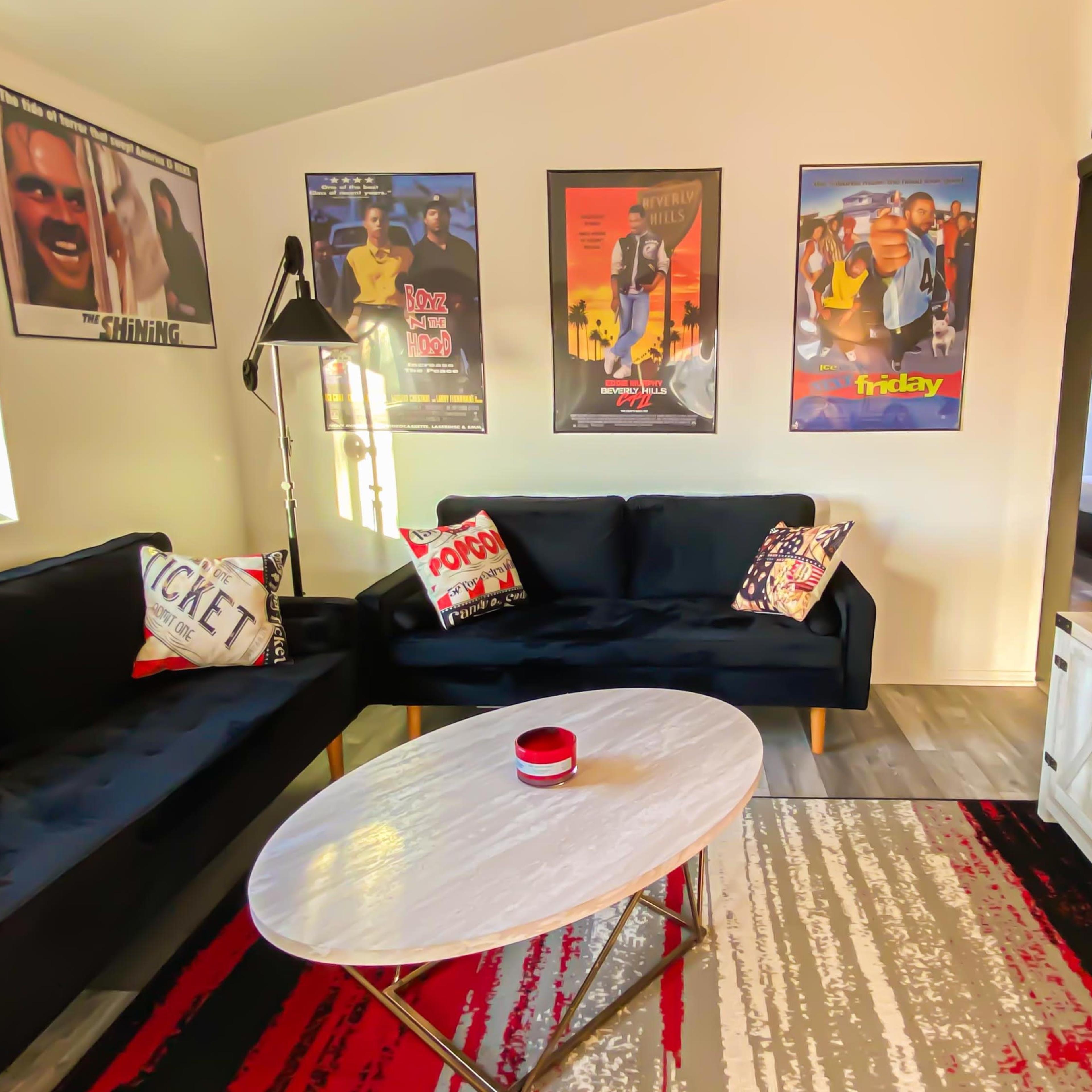 A living room with two black sofas facing a white coffee table, decorated with movie posters on the walls and a mounted television.