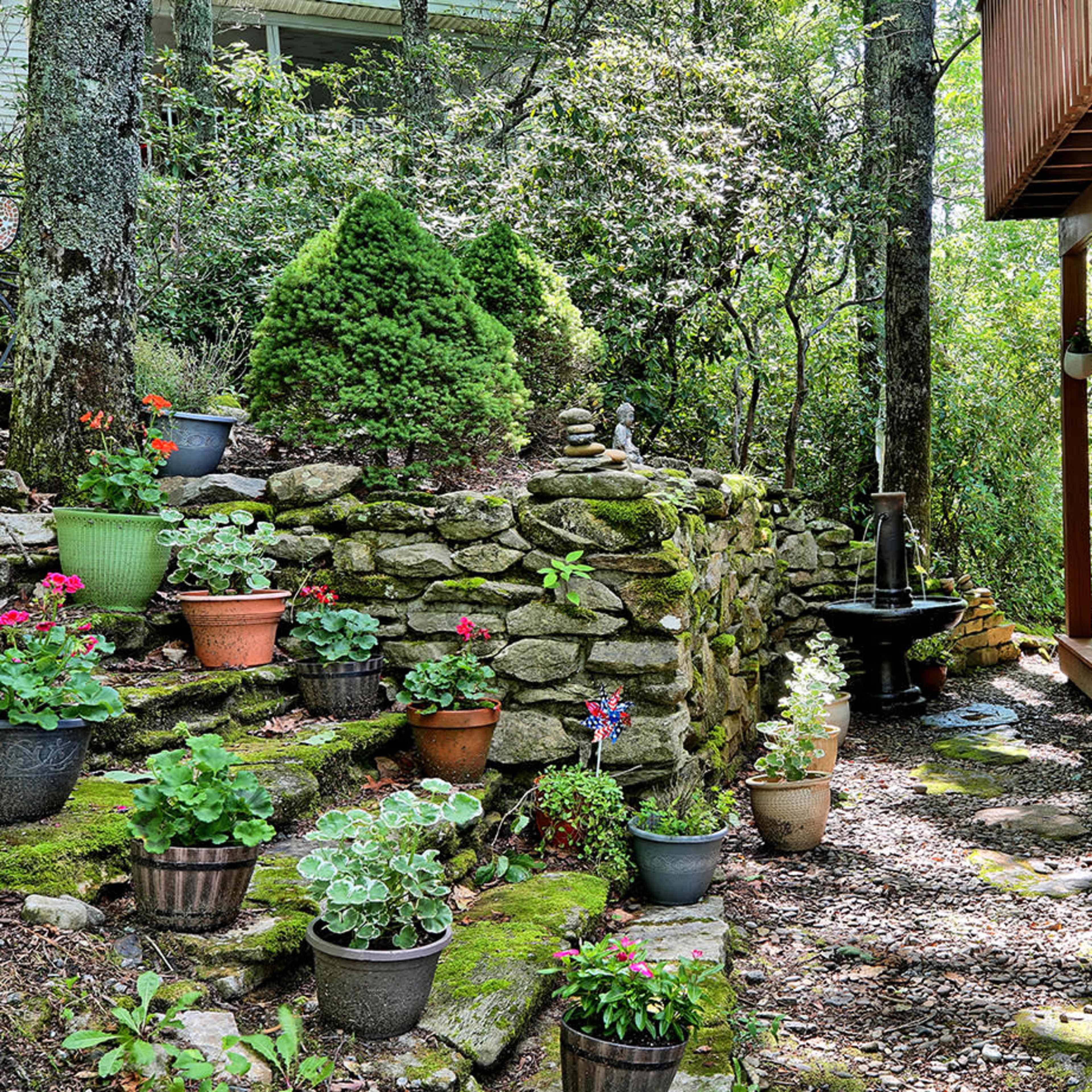 A stone wall garden path with various potted plants leads to a wooden deck surrounded by trees.