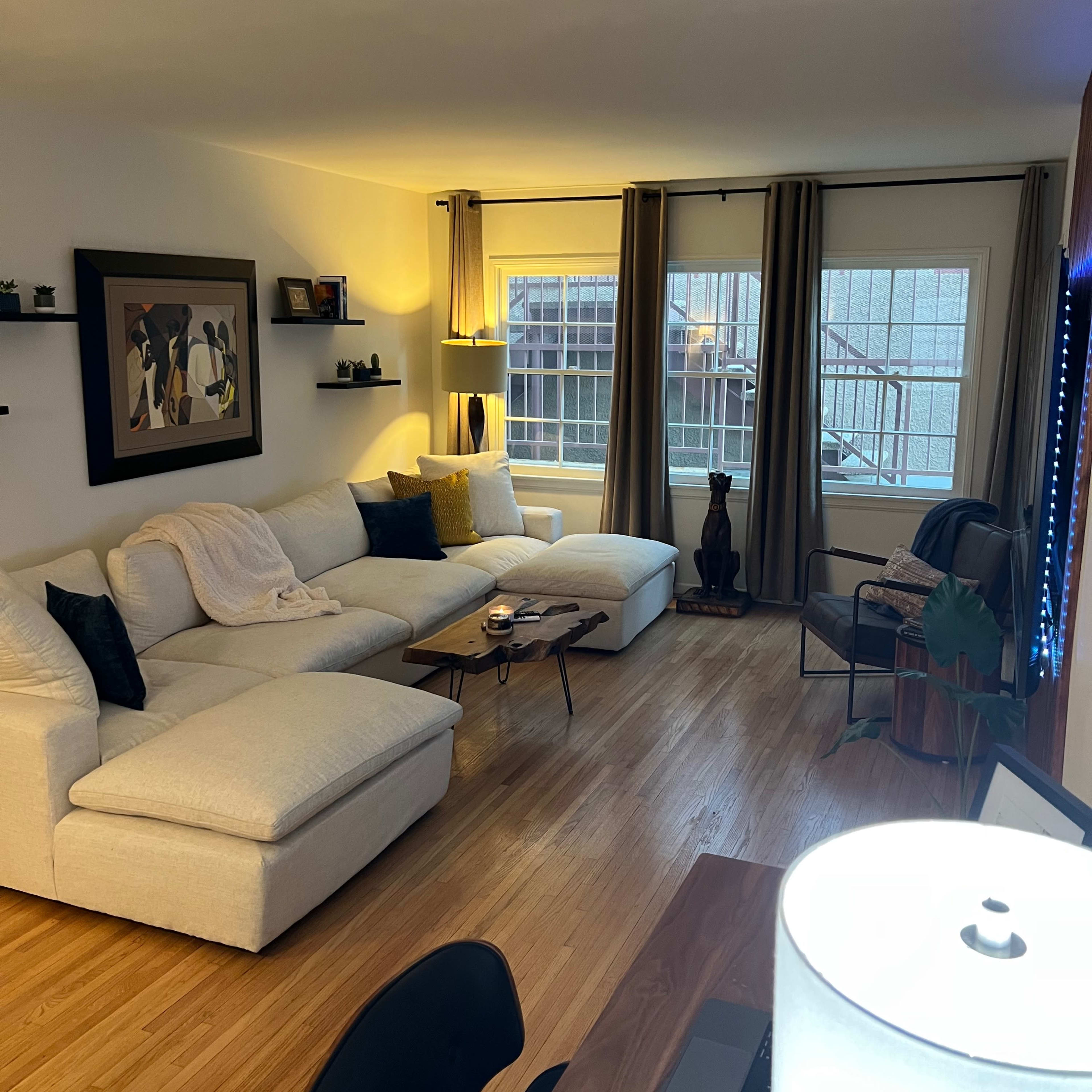 A bright living room features a large sectional sofa, a coffee table, and a desk, with large windows dressed in curtains.