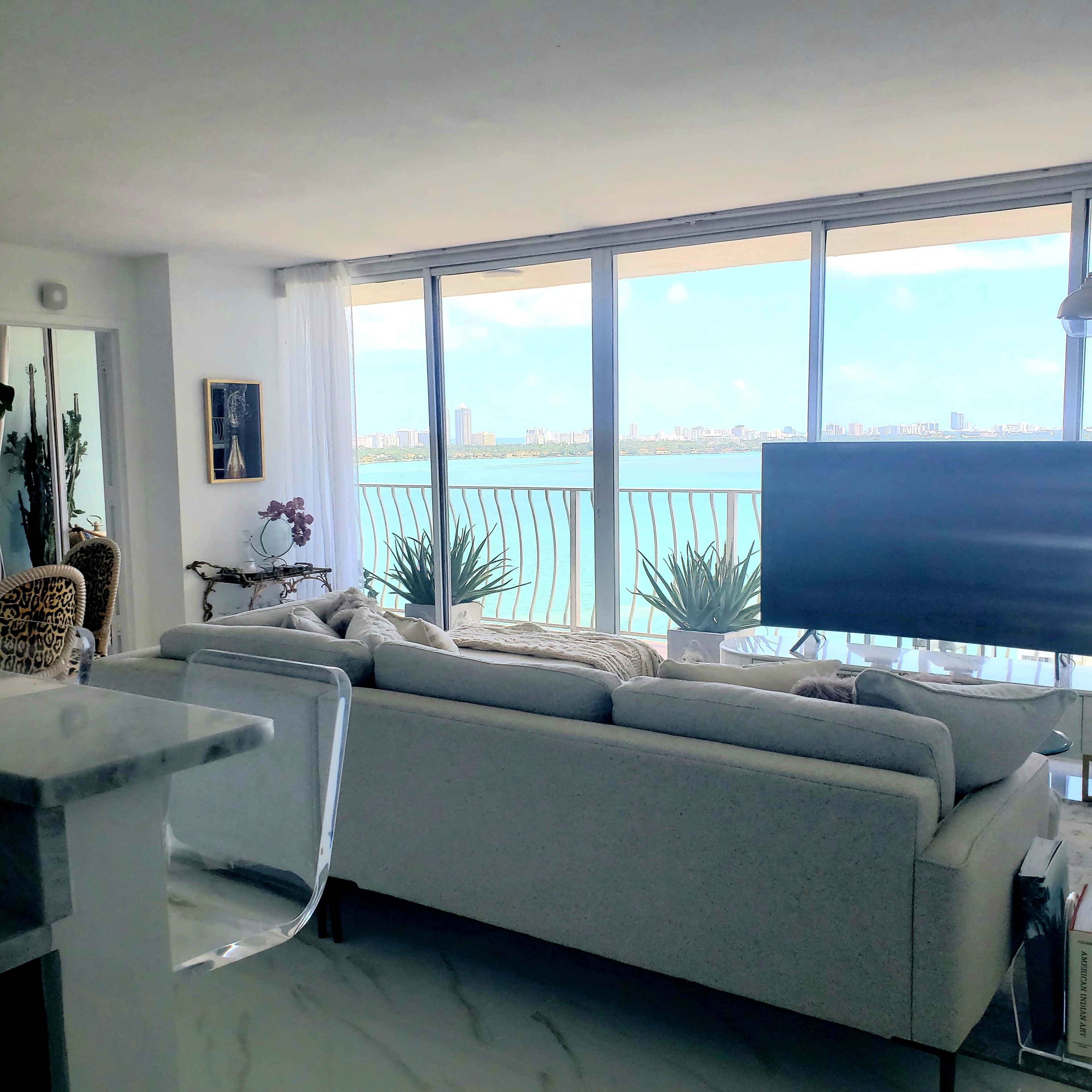 A modern living room features a white sofa facing large windows with a view of a waterfront city skyline.