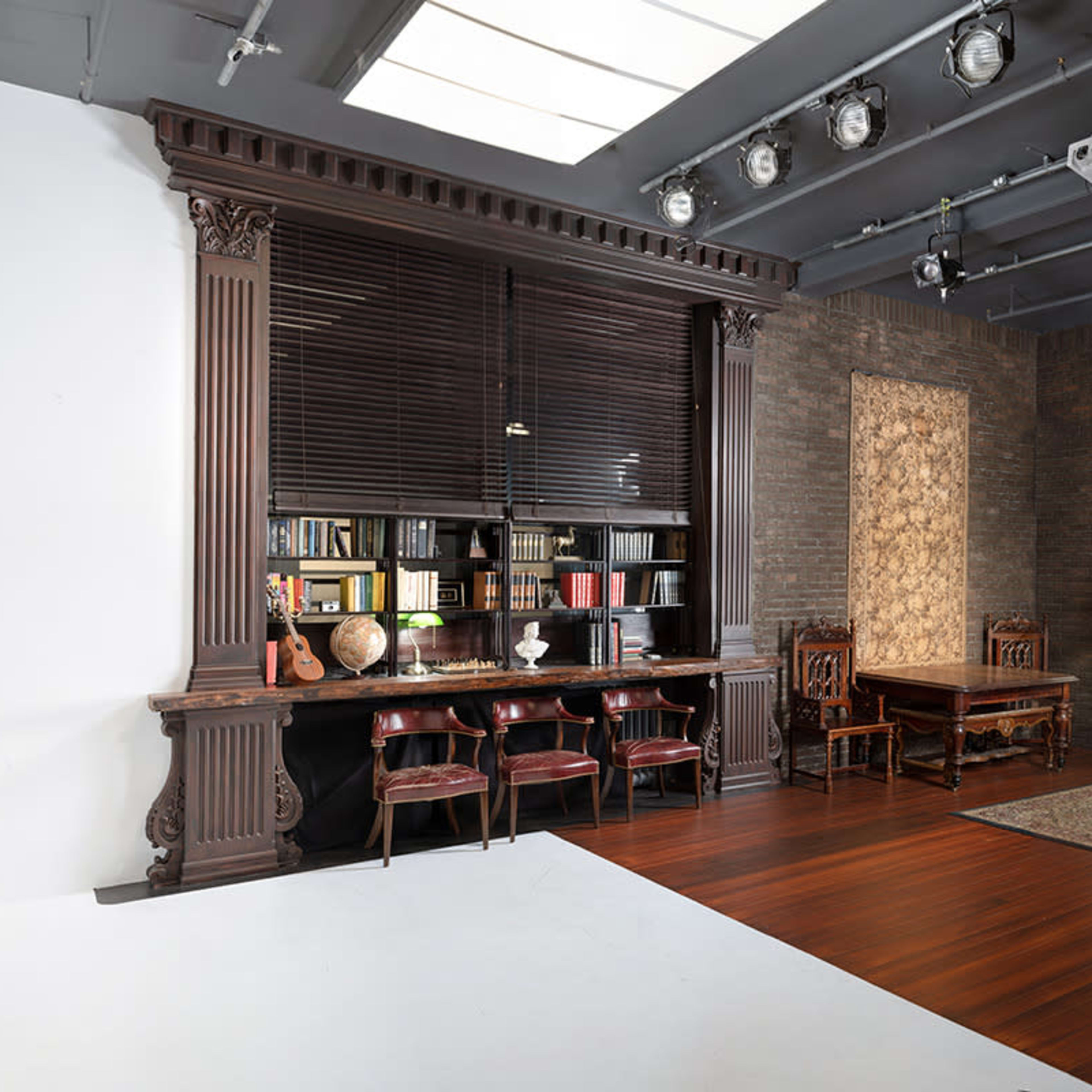 A studio setup featuring a brown leather couch, a bookshelf filled with books, a wooden desk, and decorative elements in a well-lit space with a combination of dark walls and wooden flooring.