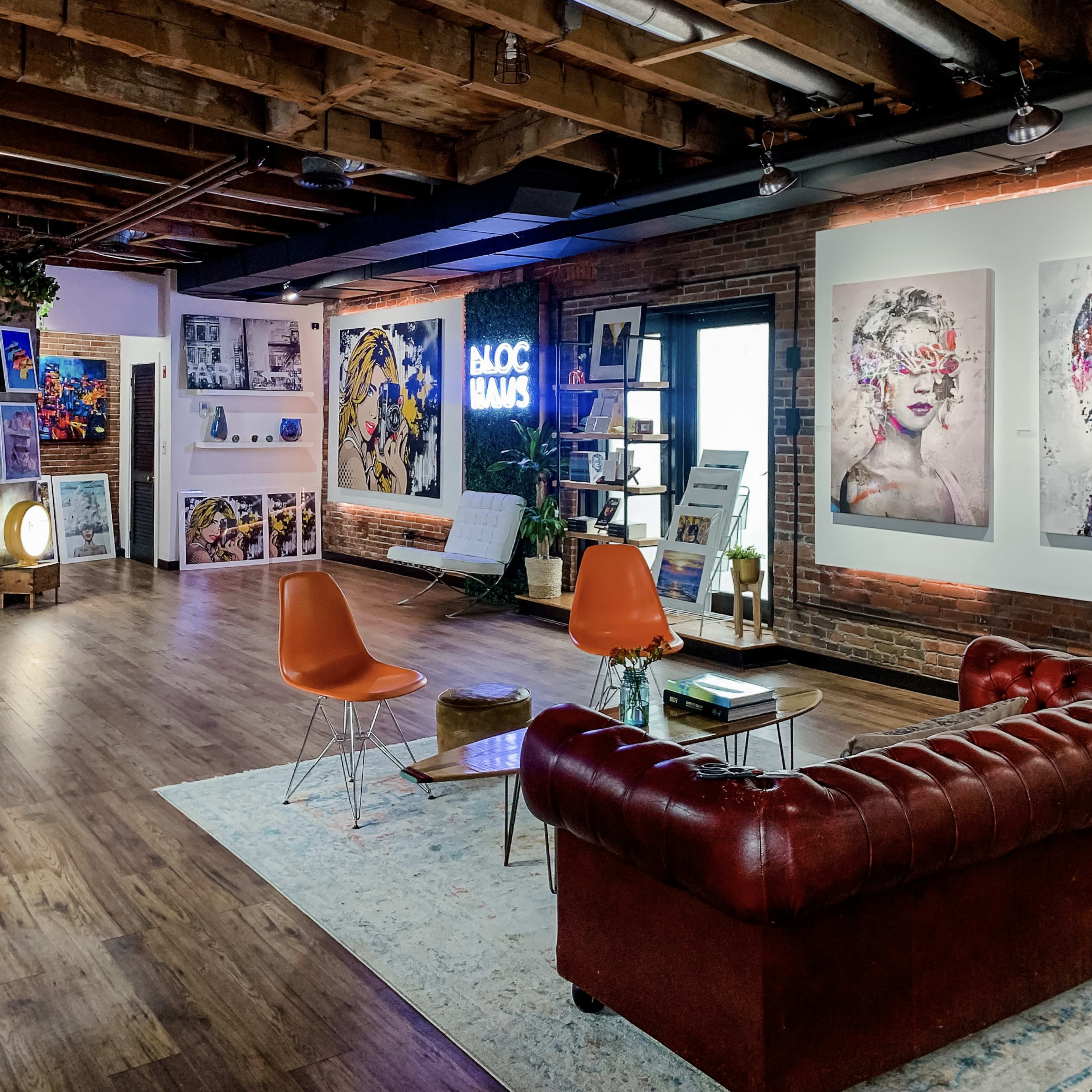A contemporary art studio featuring a mix of colorful artwork on the walls, a red leather sofa, orange chairs, and wooden beams on the ceiling.