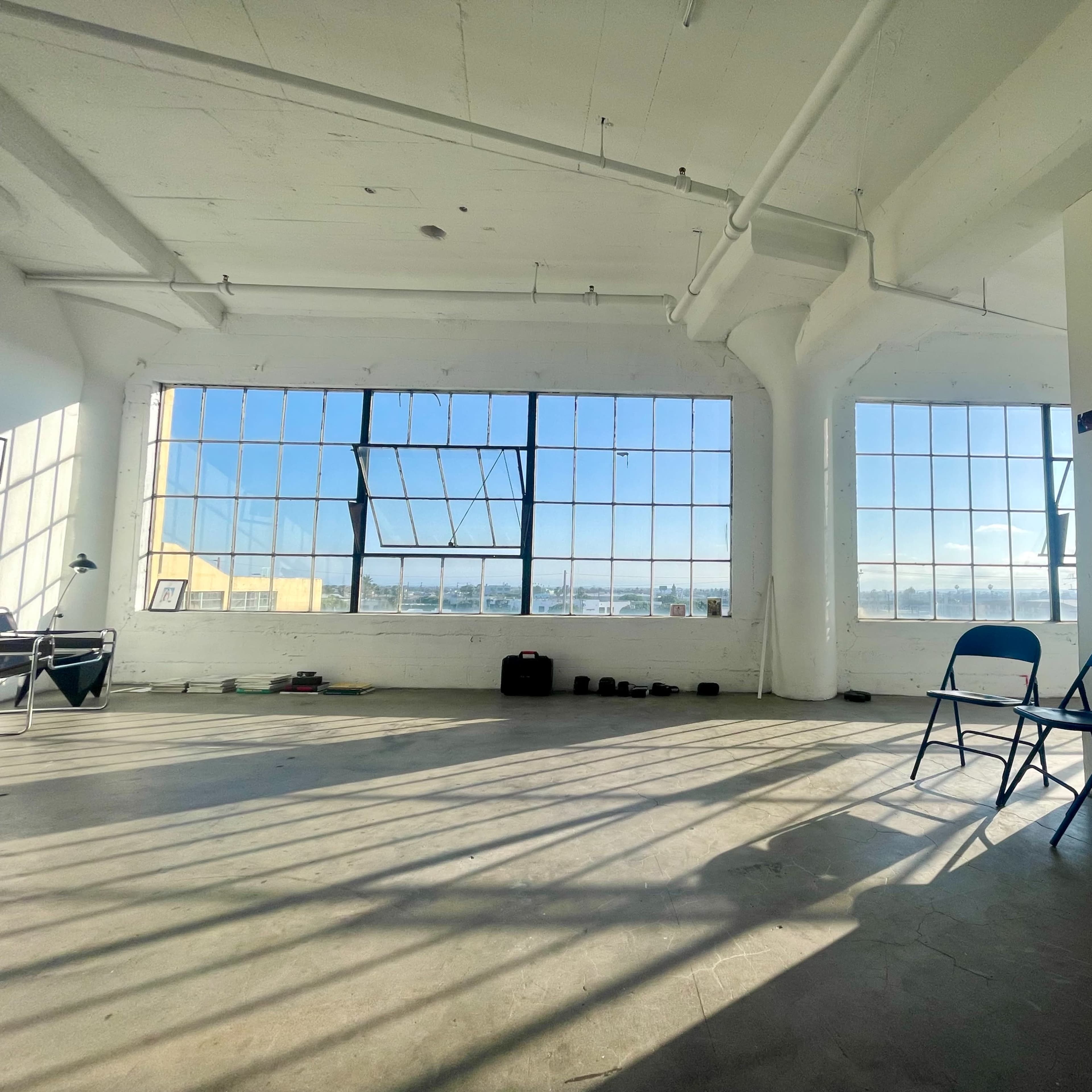 A spacious room with large windows providing sunlight, two blue chairs, and some weights on the floor.