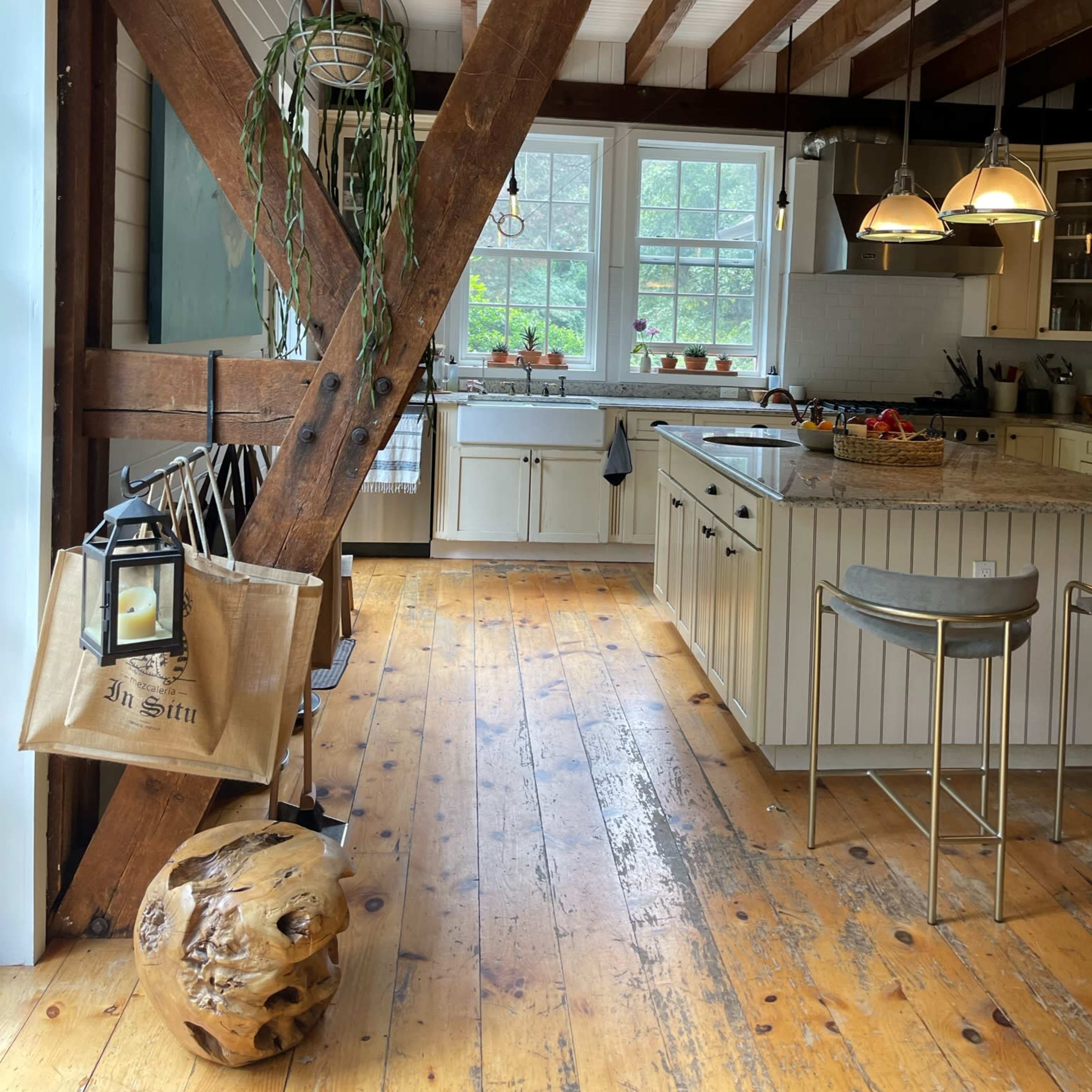 A rustic kitchen features exposed wooden beams, a central island, and large windows with greenery outside.
