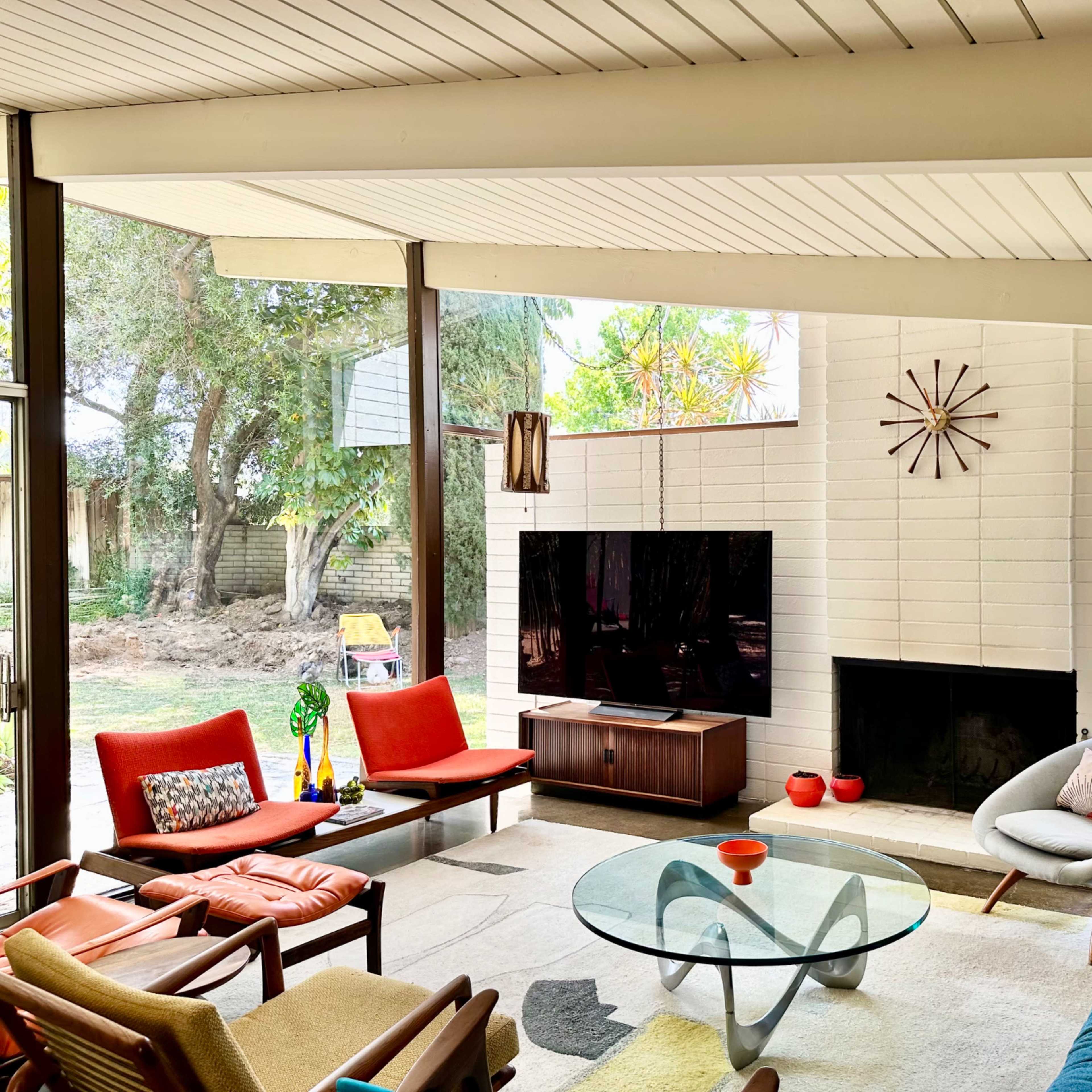 A modern living room with large glass windows, featuring a blue sofa, orange chairs, a glass coffee table, and a wall-mounted television above a white fireplace.
