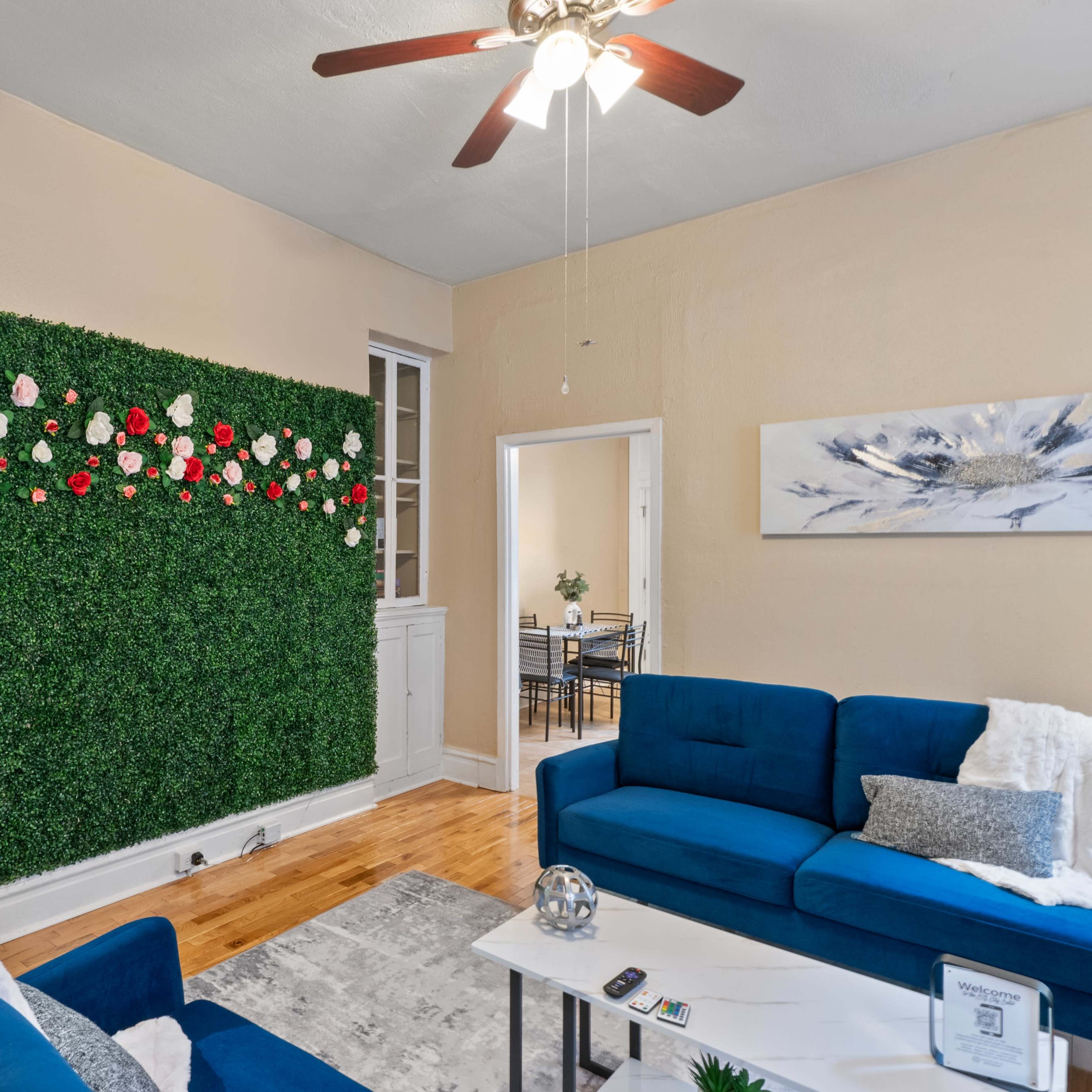 The living room features a green wall adorned with artificial flowers, flanked by two blue sofas and a light-colored rug.