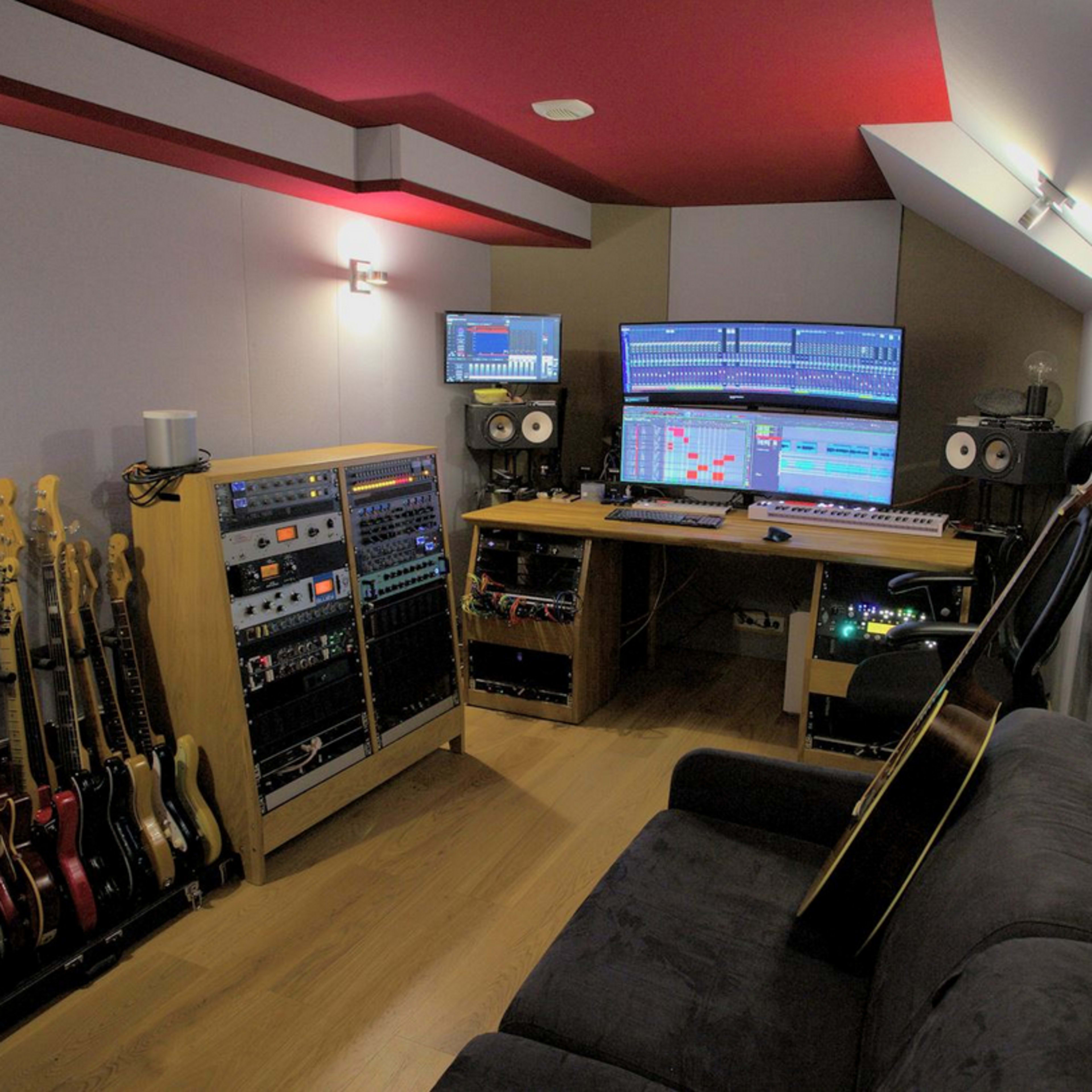 A music production studio features multiple guitars on stands, audio equipment, and dual computer monitors displaying audio software, all arranged in a compact, modern space.