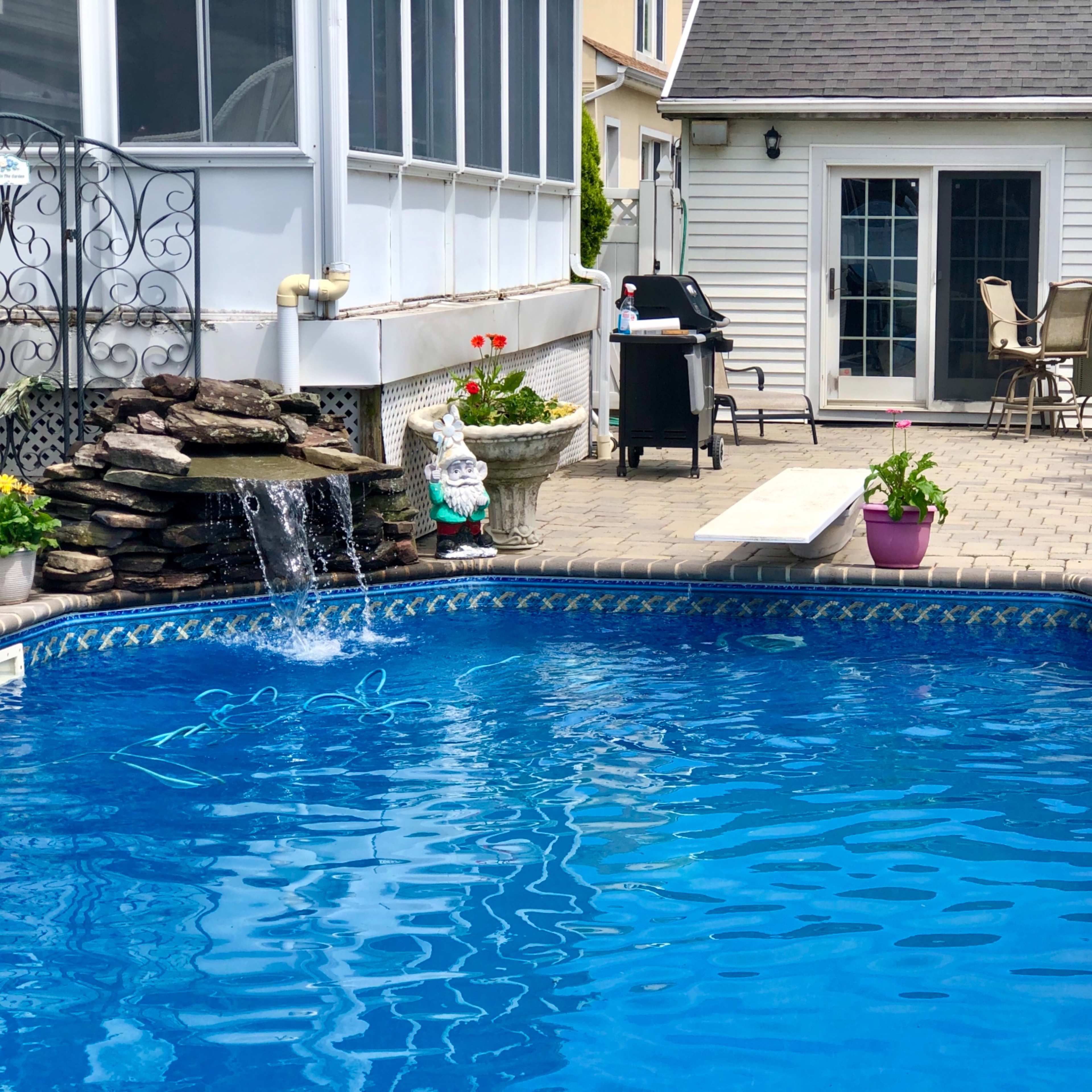 A clear blue swimming pool is surrounded by potted plants, outdoor furniture, and a nearby stone waterfall.