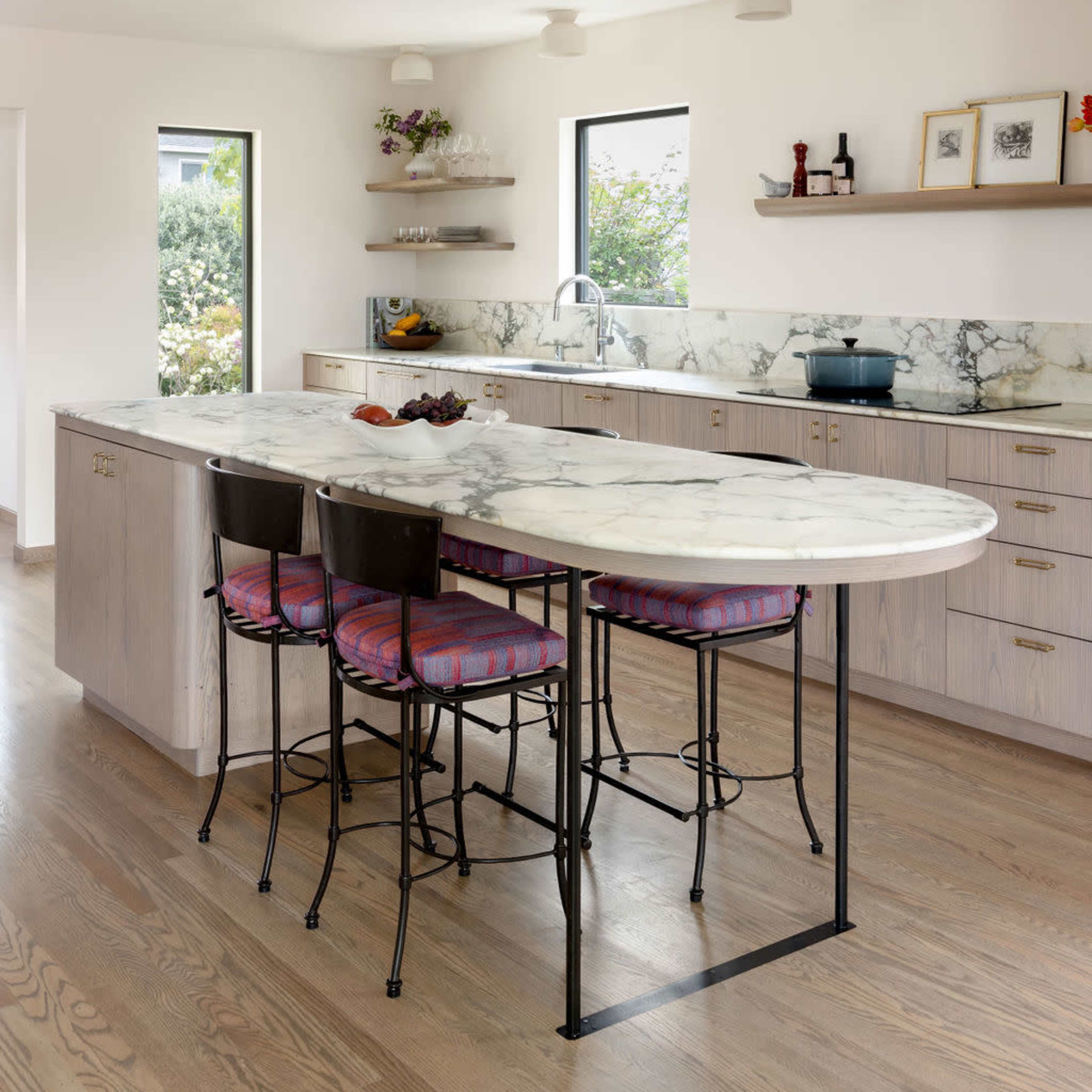 A modern kitchen features a curved marble countertop with four high stools, light wood cabinetry, and large windows allowing natural light.