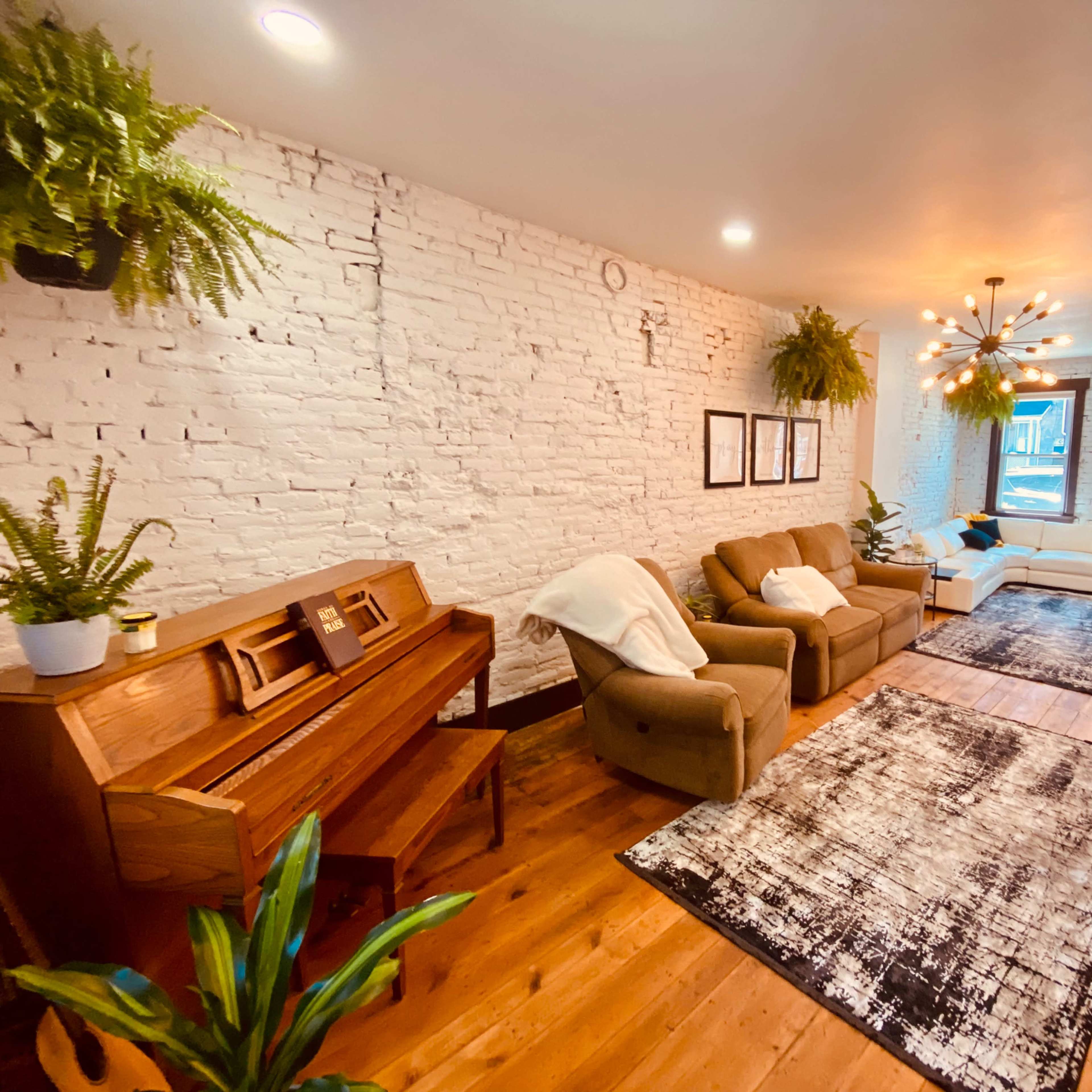A spacious living room with white brick walls, wooden flooring, two couches, a piano, and plants hanging from the ceiling.