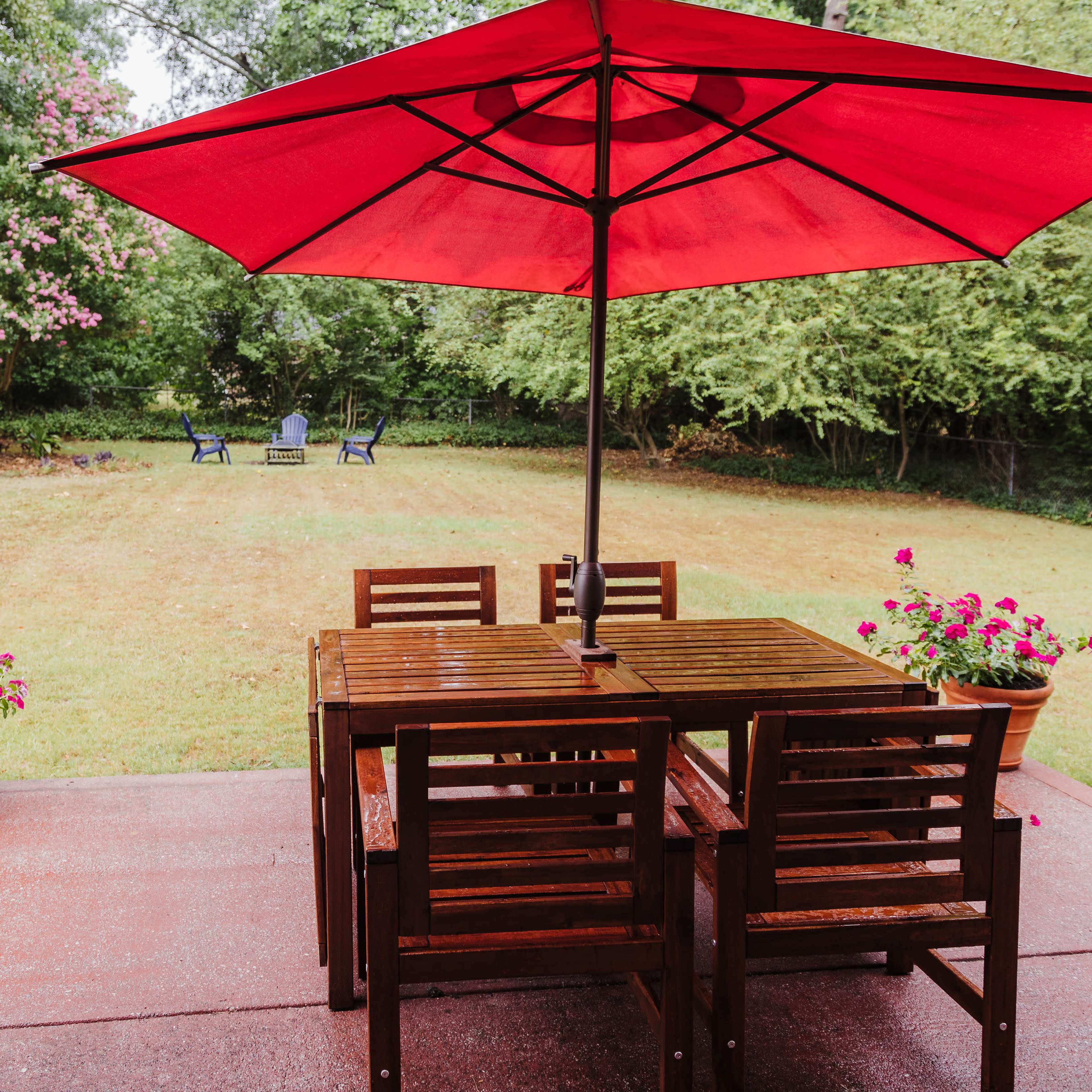 A wooden patio table with chairs is shaded by a red umbrella, surrounded by a grassy backyard and flower pots.