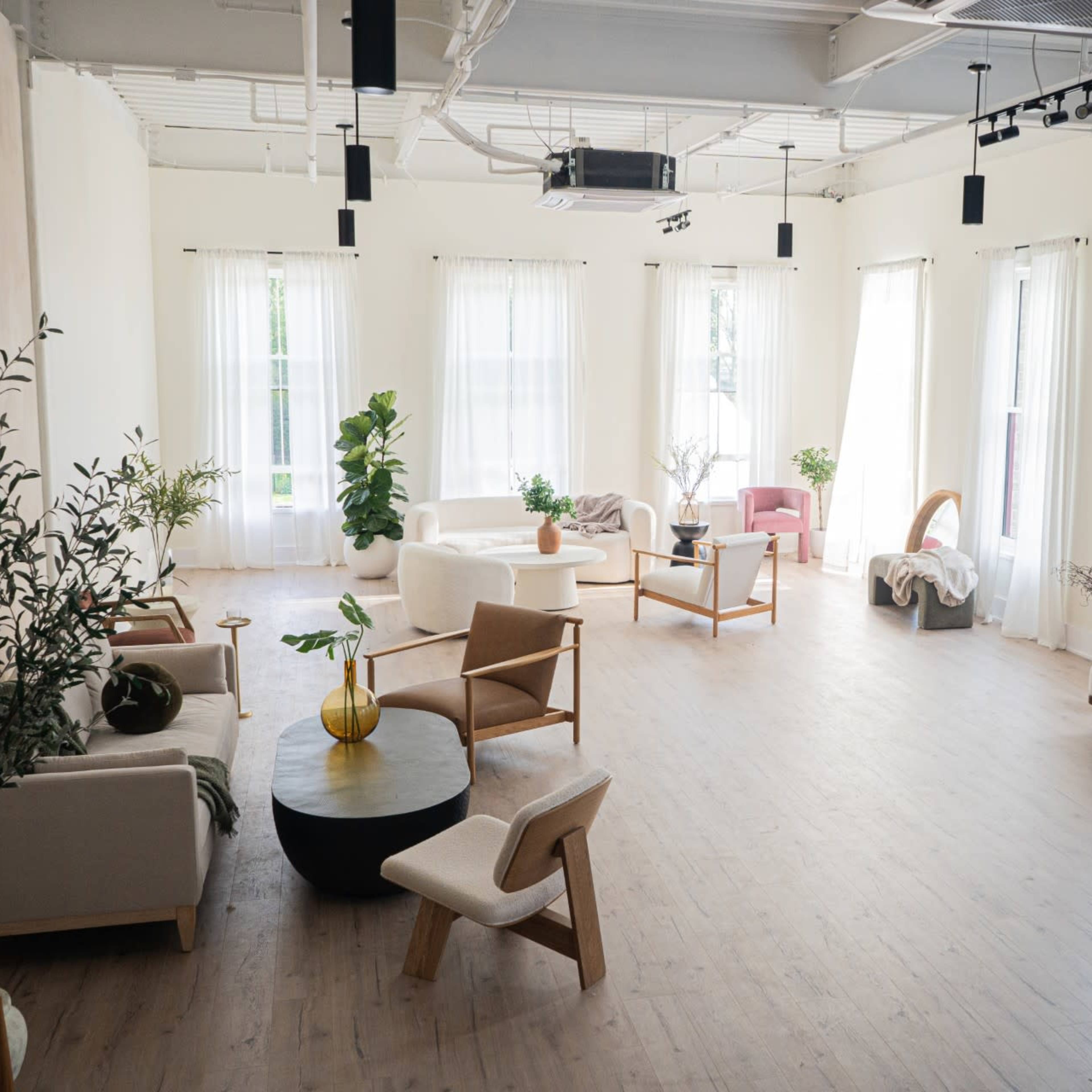 A spacious, well-lit room features several modern seating arrangements amid tall windows and decorative plants.