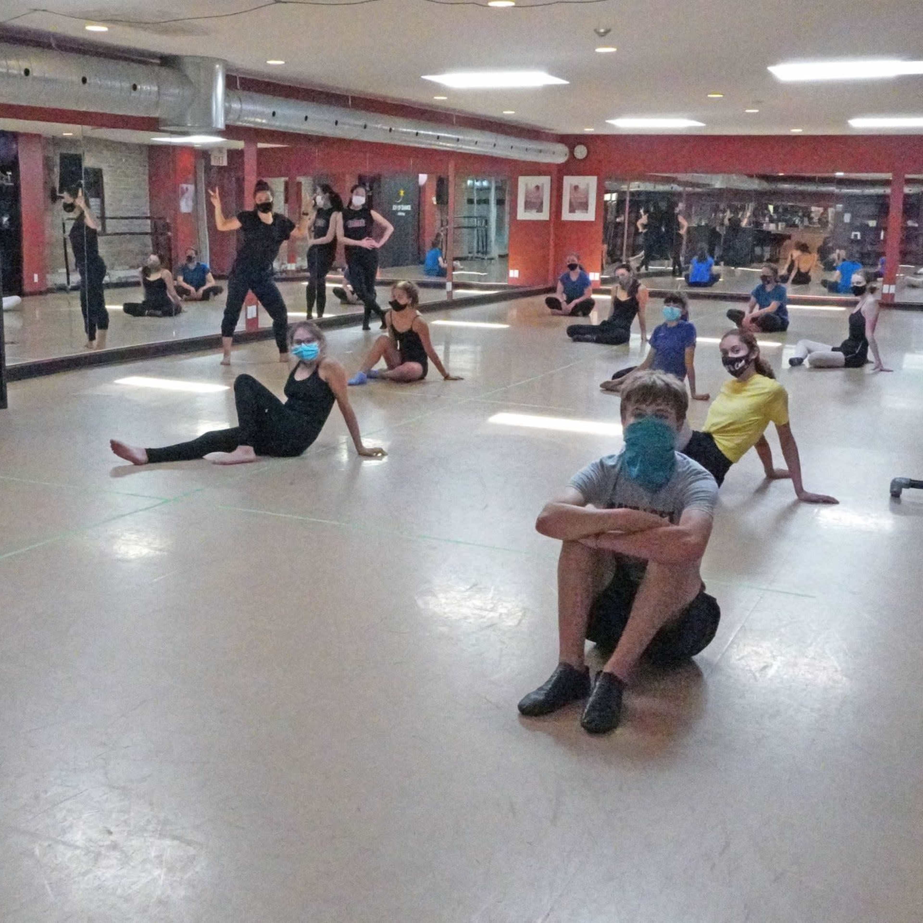 A group of dancers wearing masks is seated on the floor of a studio, with some standing and stretching in the background.