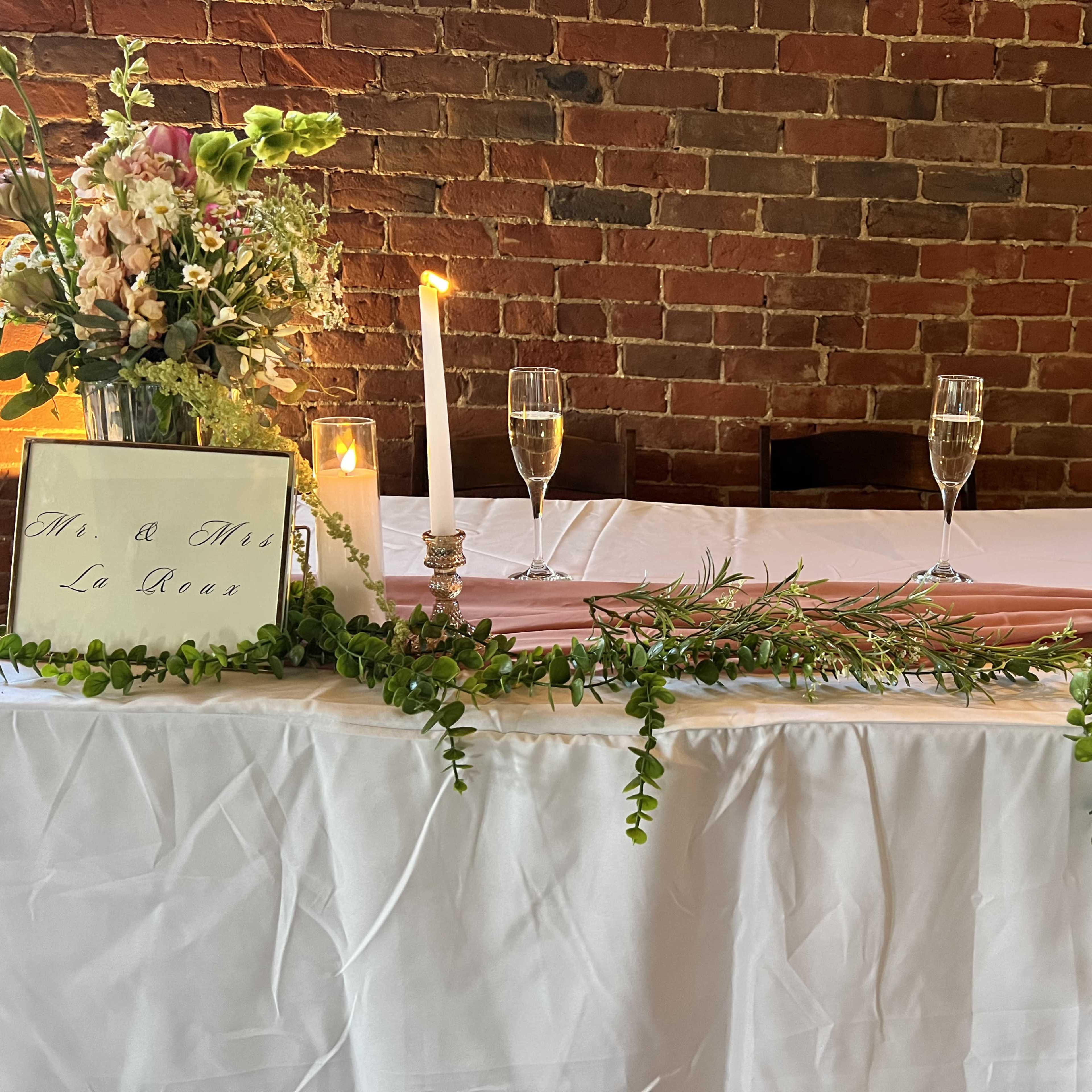 A decorated table featuring floral arrangements, candles, and champagne glasses is set against a brick wall.