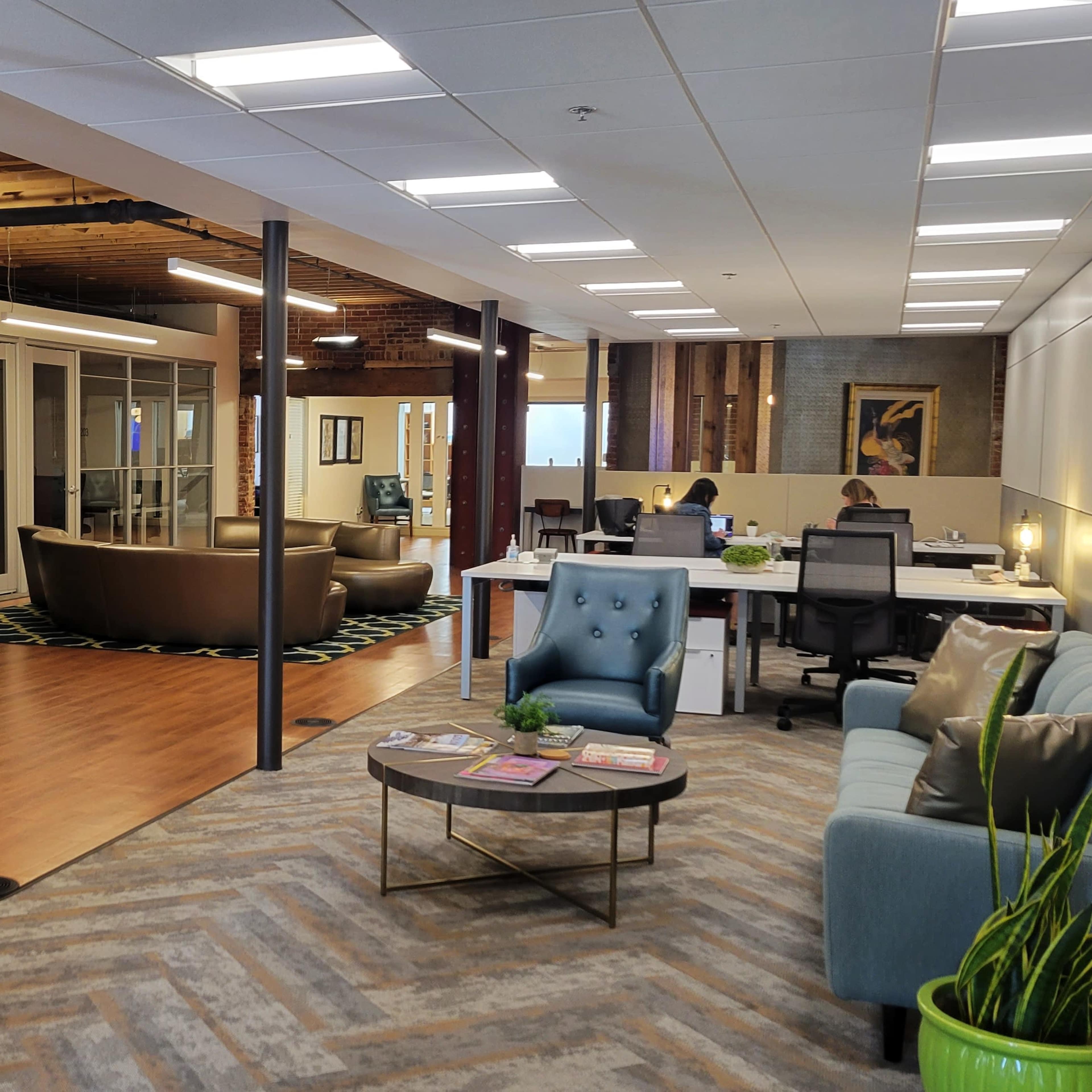 A modern office space featuring a seating area with chairs and a sofa, workstations in the background, and large glass doors leading to another section.