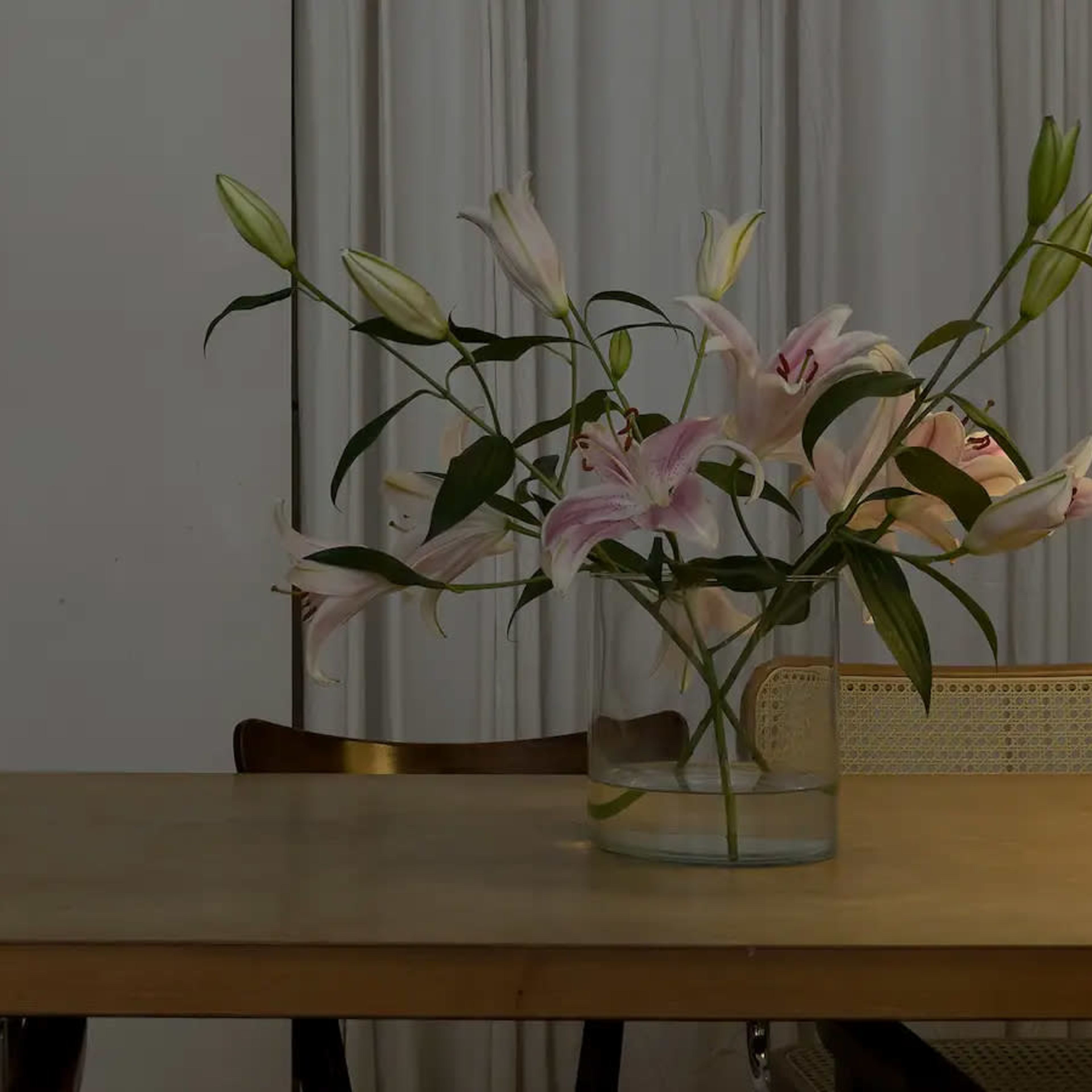 A glass vase with pink lilies is placed on a wooden table in a room featuring a curtain and a door.