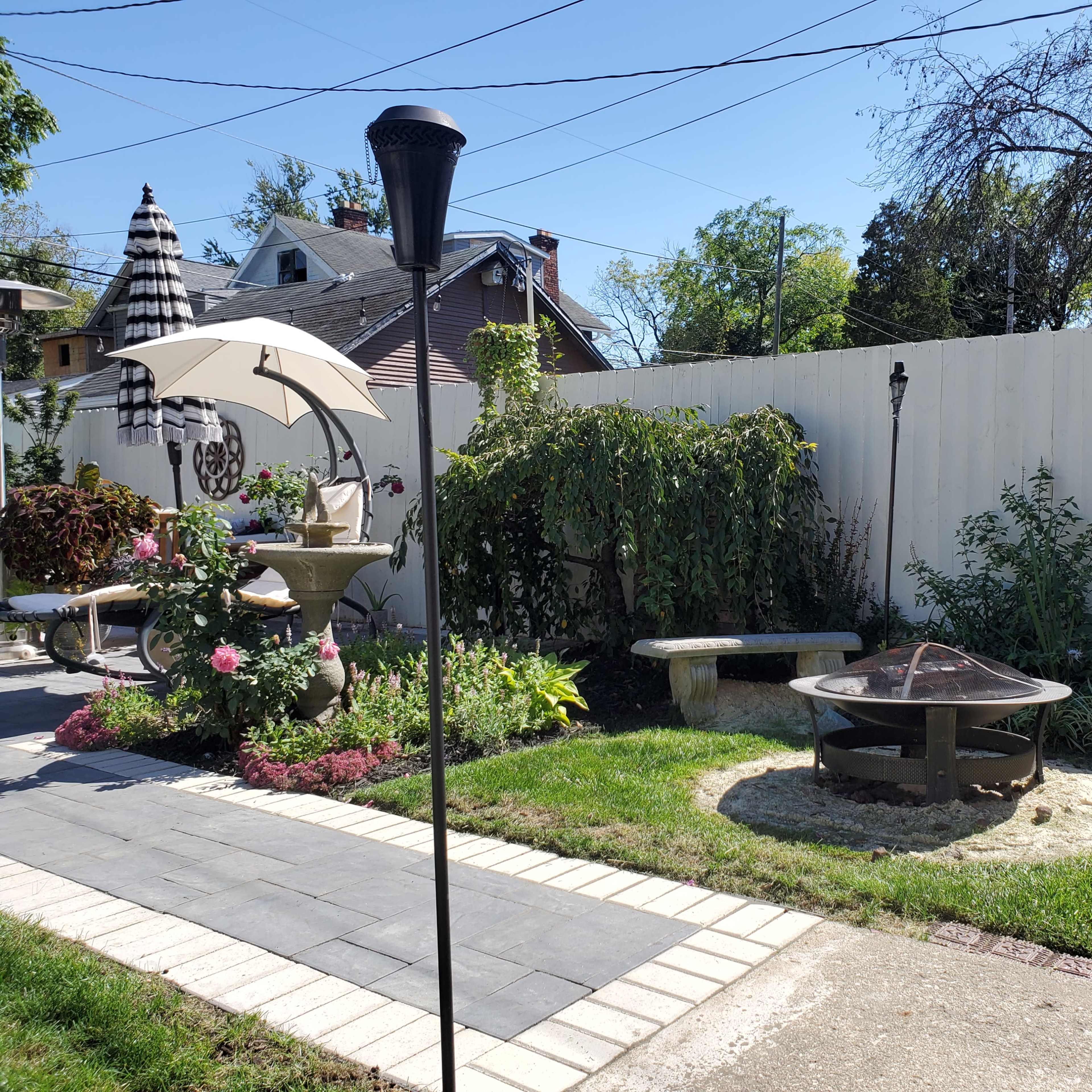 A landscaped backyard featuring a fire pit surrounded by stone benches, a patio area with an umbrella, and various plants along a white fence.