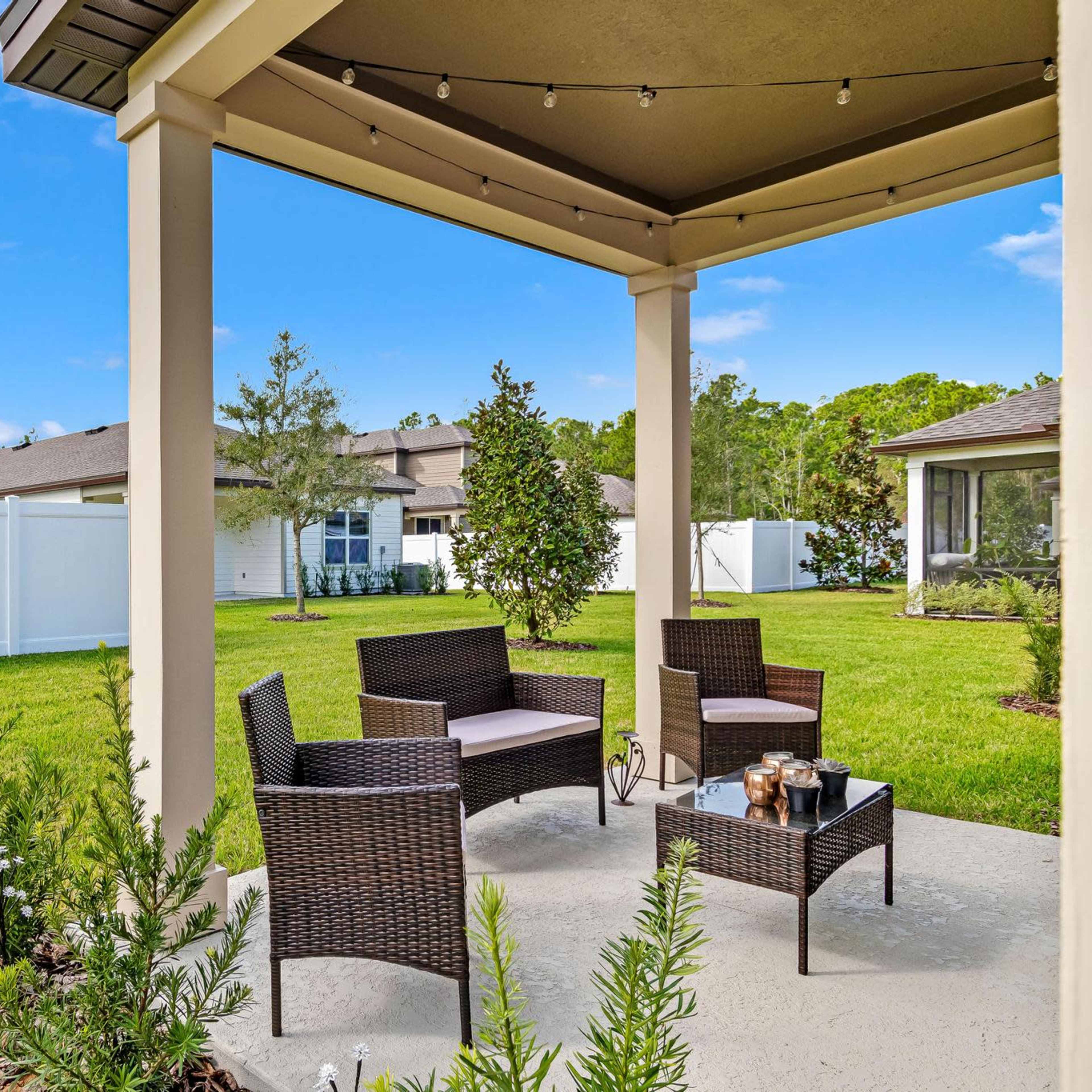 A covered patio features wicker furniture set around a small table in a backyard with a neat lawn and white privacy fence.