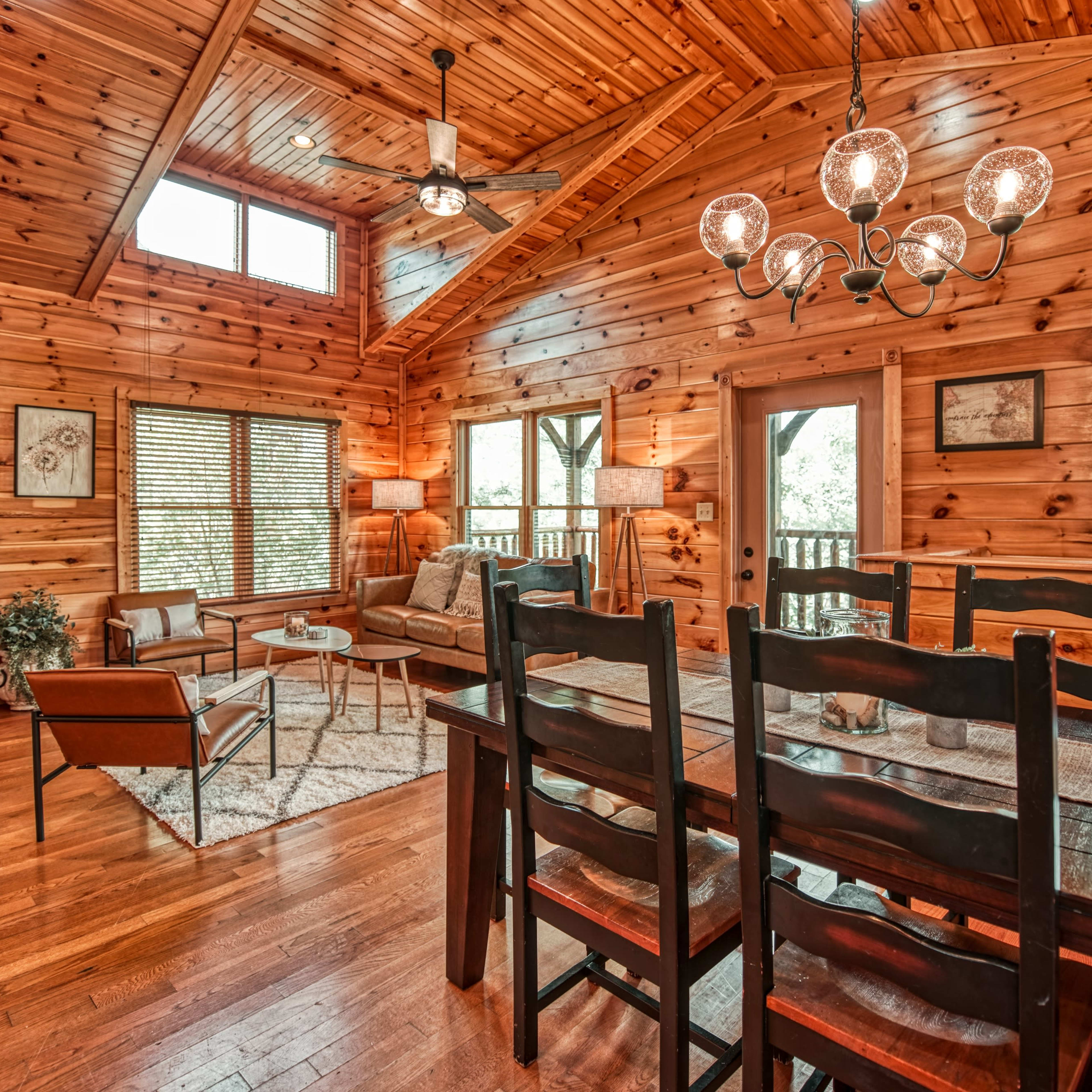 The interior of a wooden cabin features a dining table with black chairs, a stone fireplace, and large windows that allow natural light to fill the open living space.