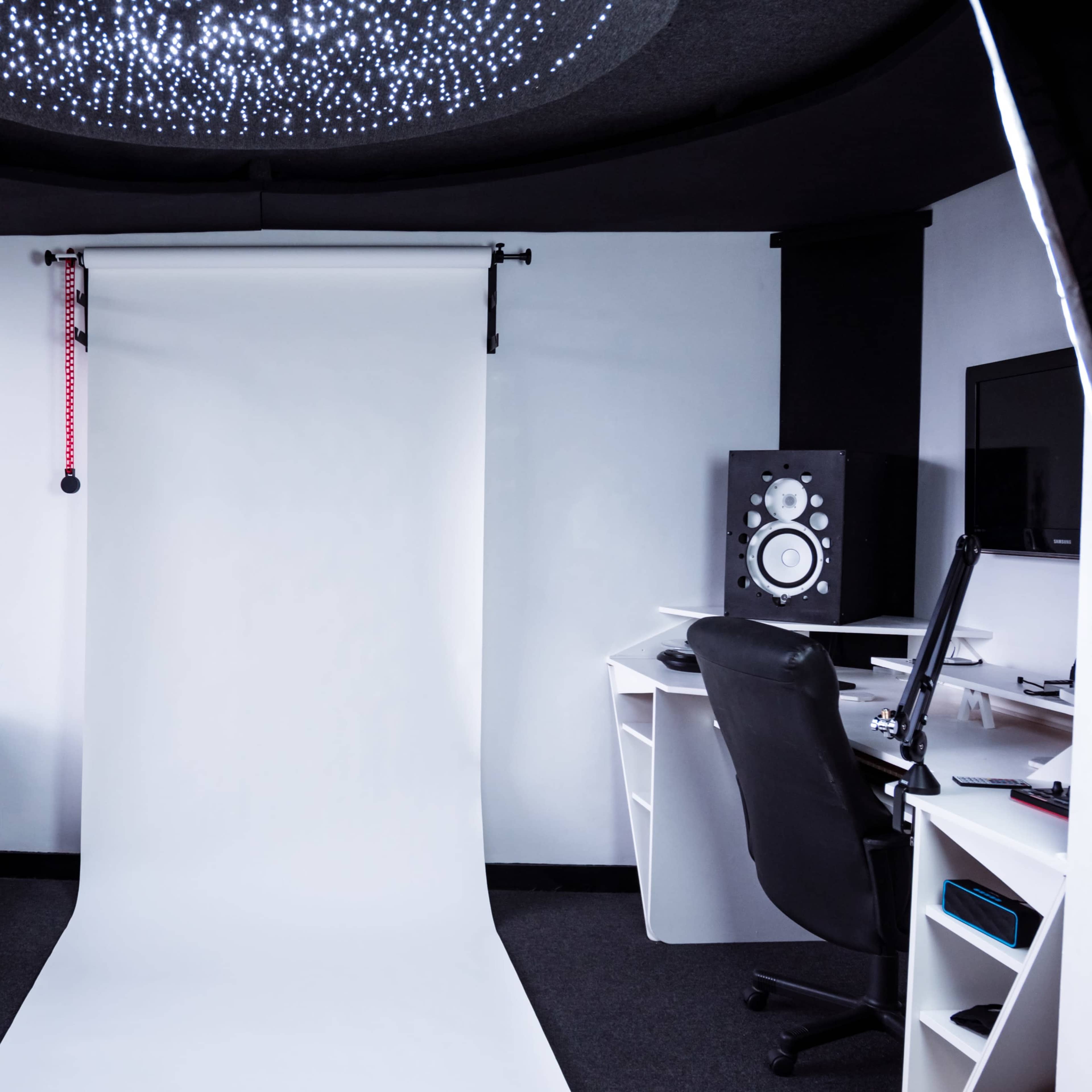 A minimalist studio space features a white backdrop, a black couch, and a desk with audio equipment and a computer.