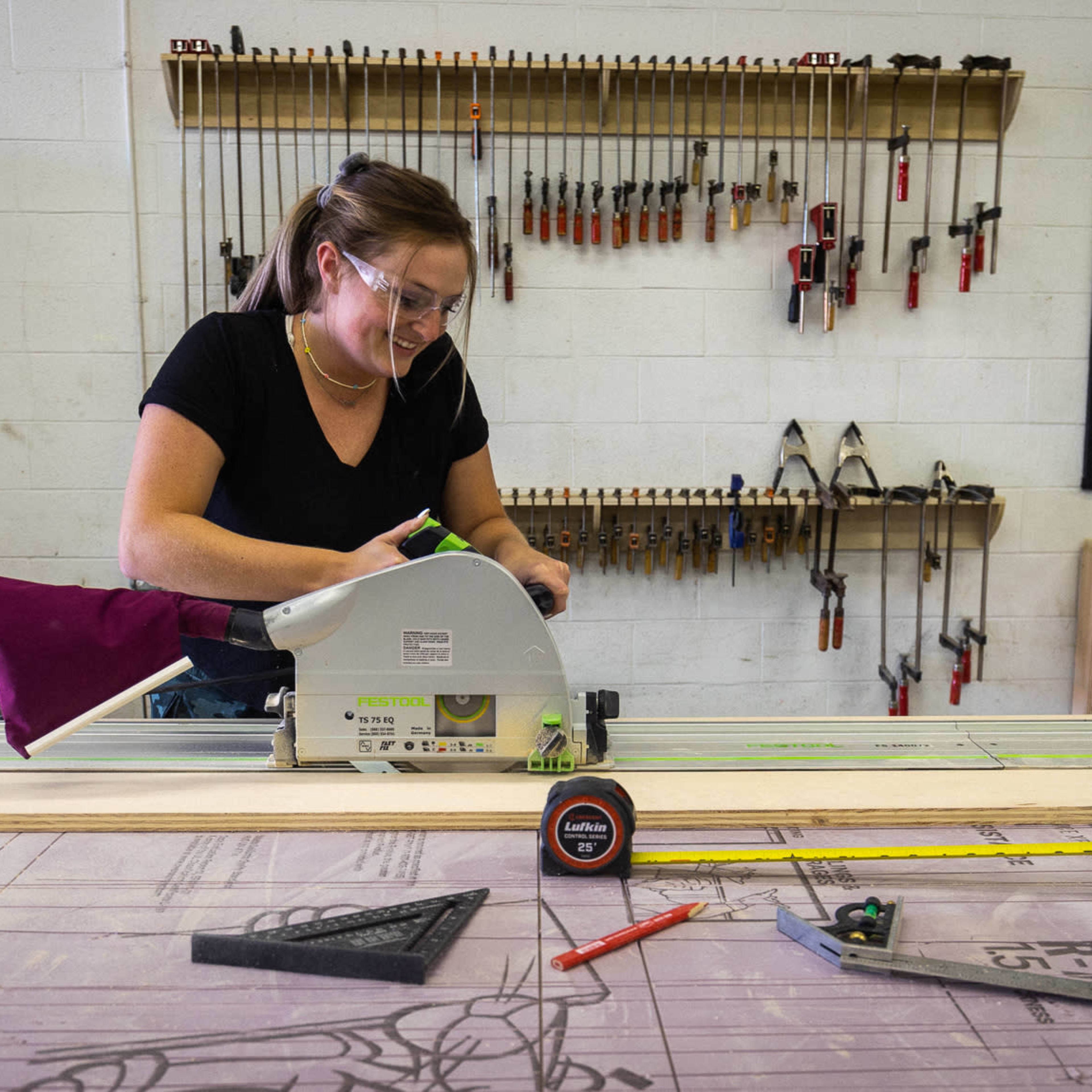 A woman operates a circular saw on a workbench in a well-organized workshop filled with various tools.