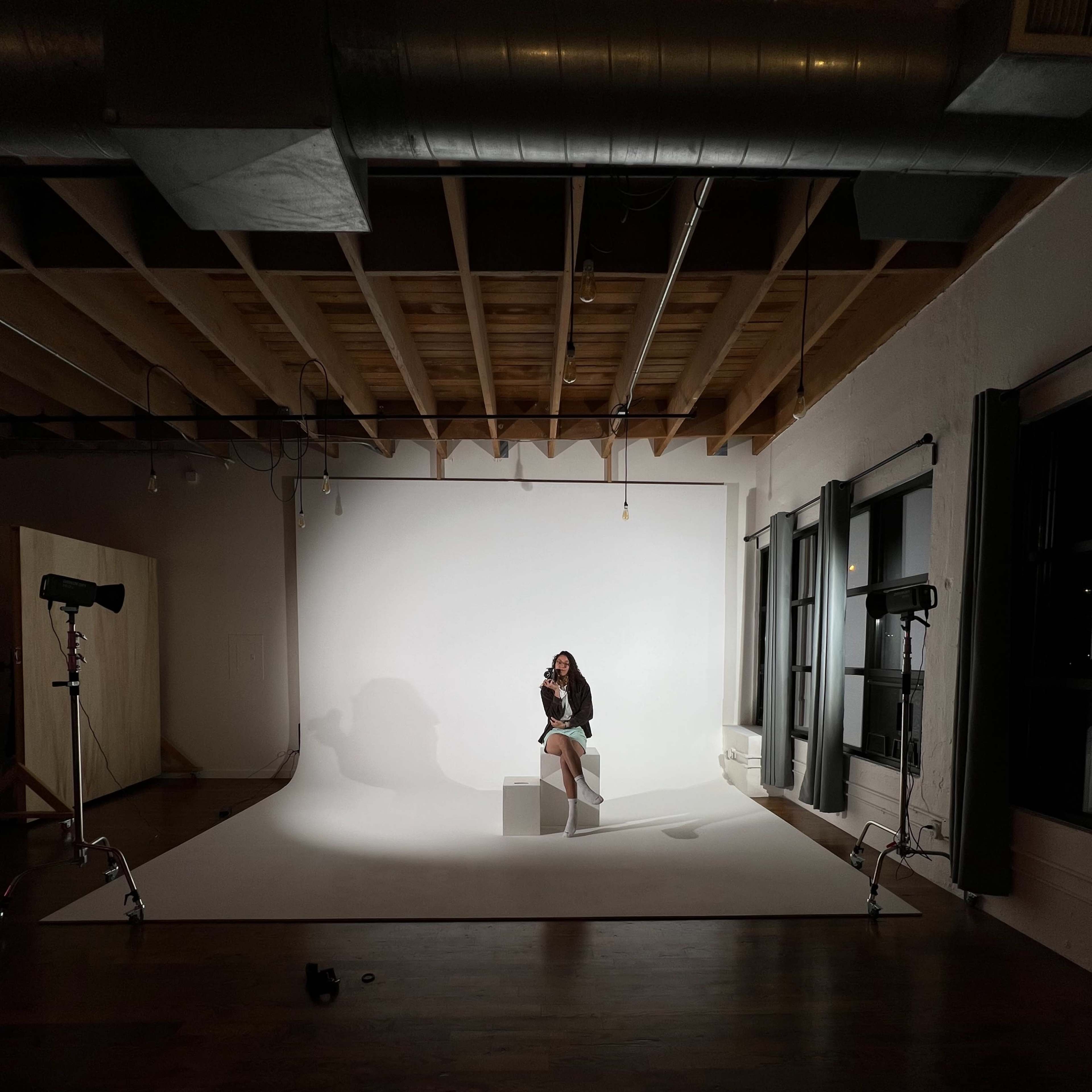 A model sits on a stool in front of a white backdrop, surrounded by camera equipment in a spacious studio.