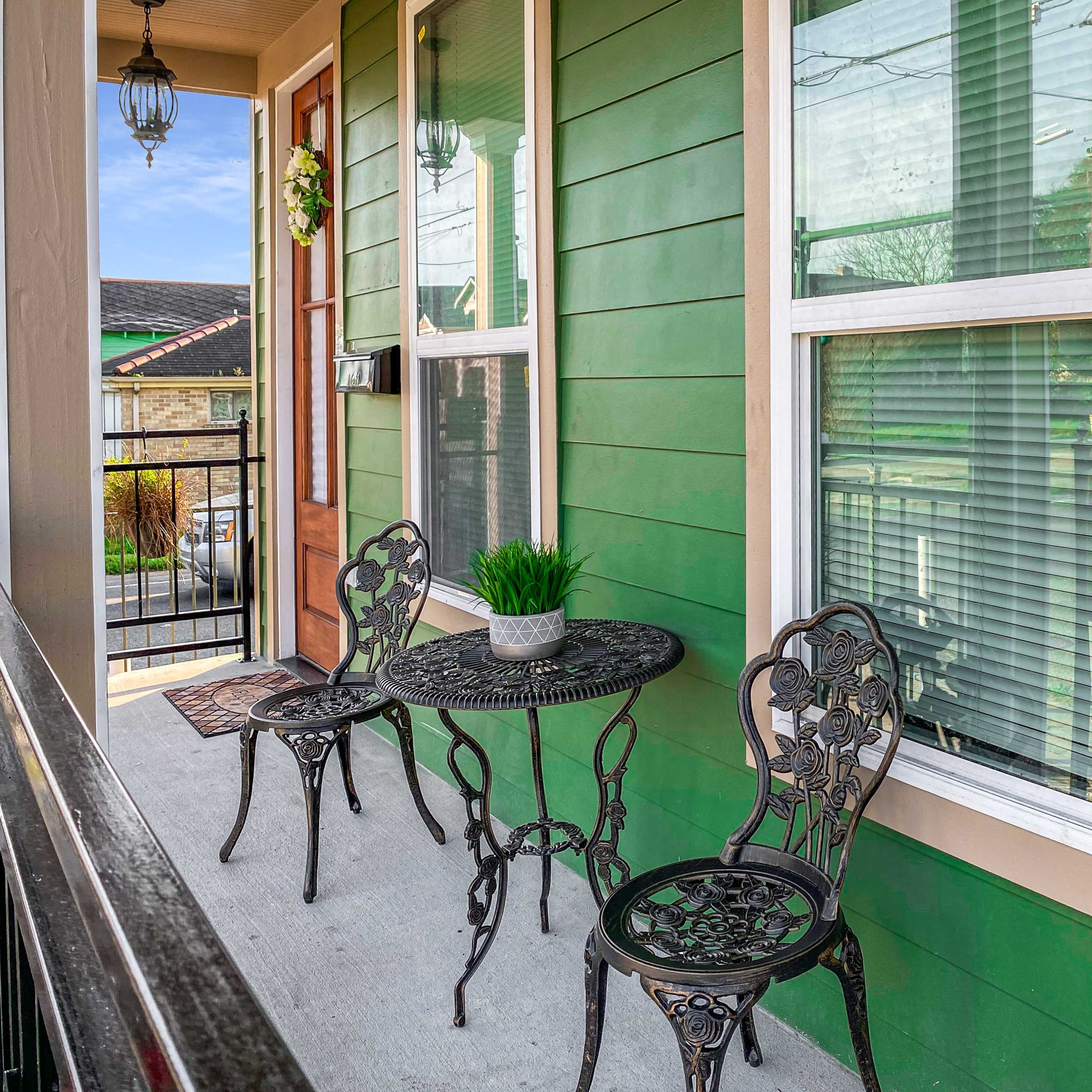 A small porch with a green exterior, featuring a round table and two metal chairs, and decorative plants near the windows.
