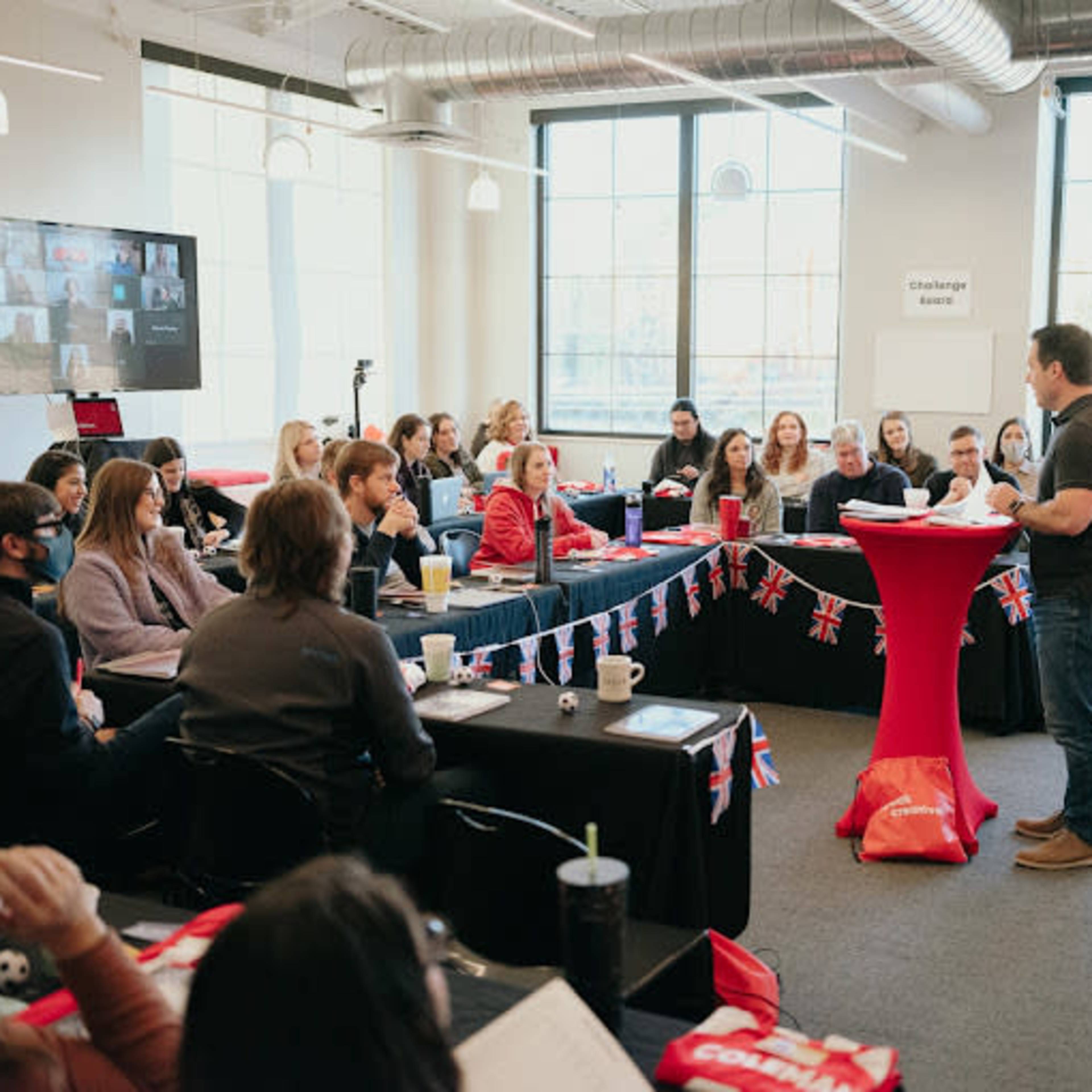 A speaker stands in front of an audience in a classroom setting, with participants seated at tables decorated with British flags.