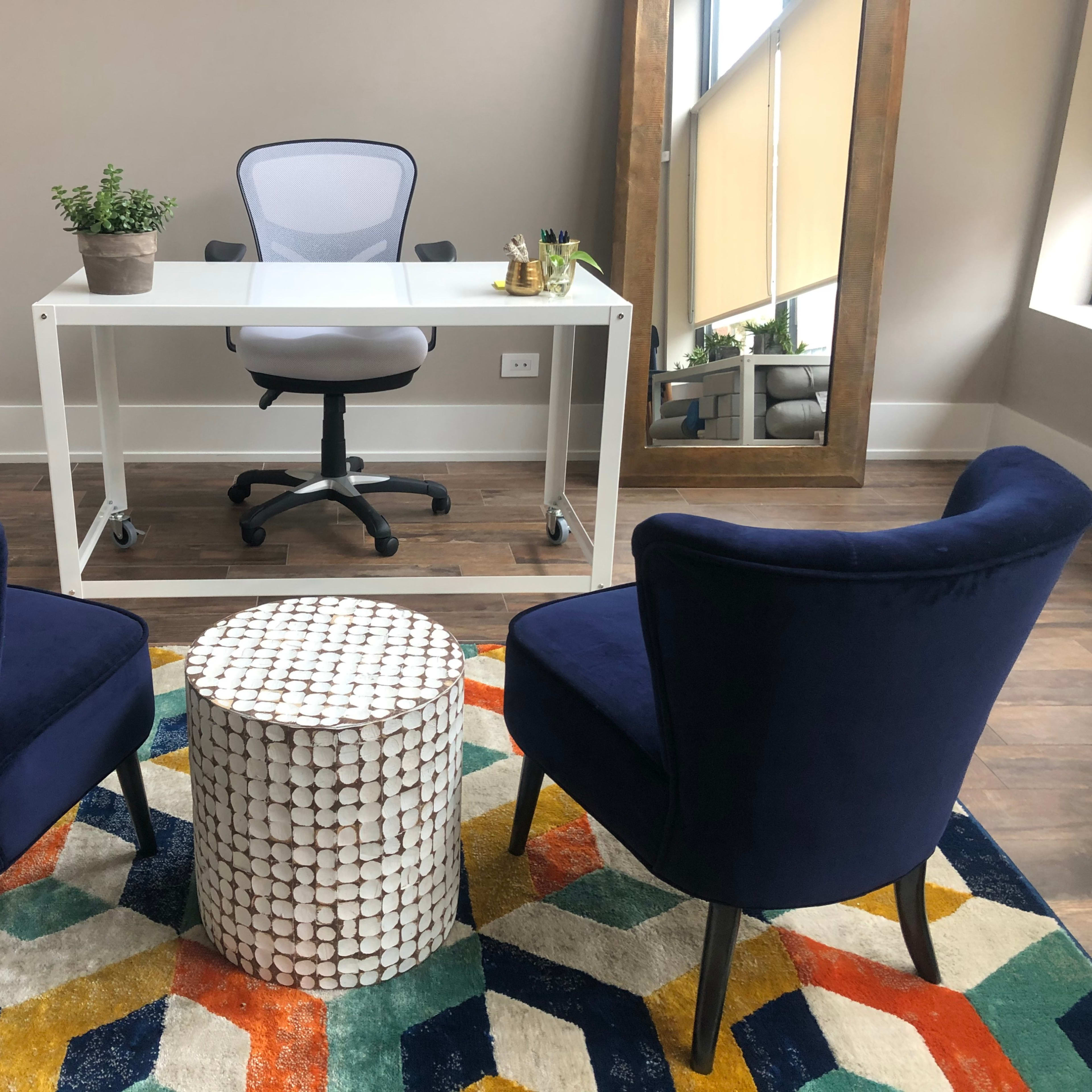 A modern office space featuring a white desk with a chair, two blue upholstered chairs, and a round, patterned table on a colorful rug.