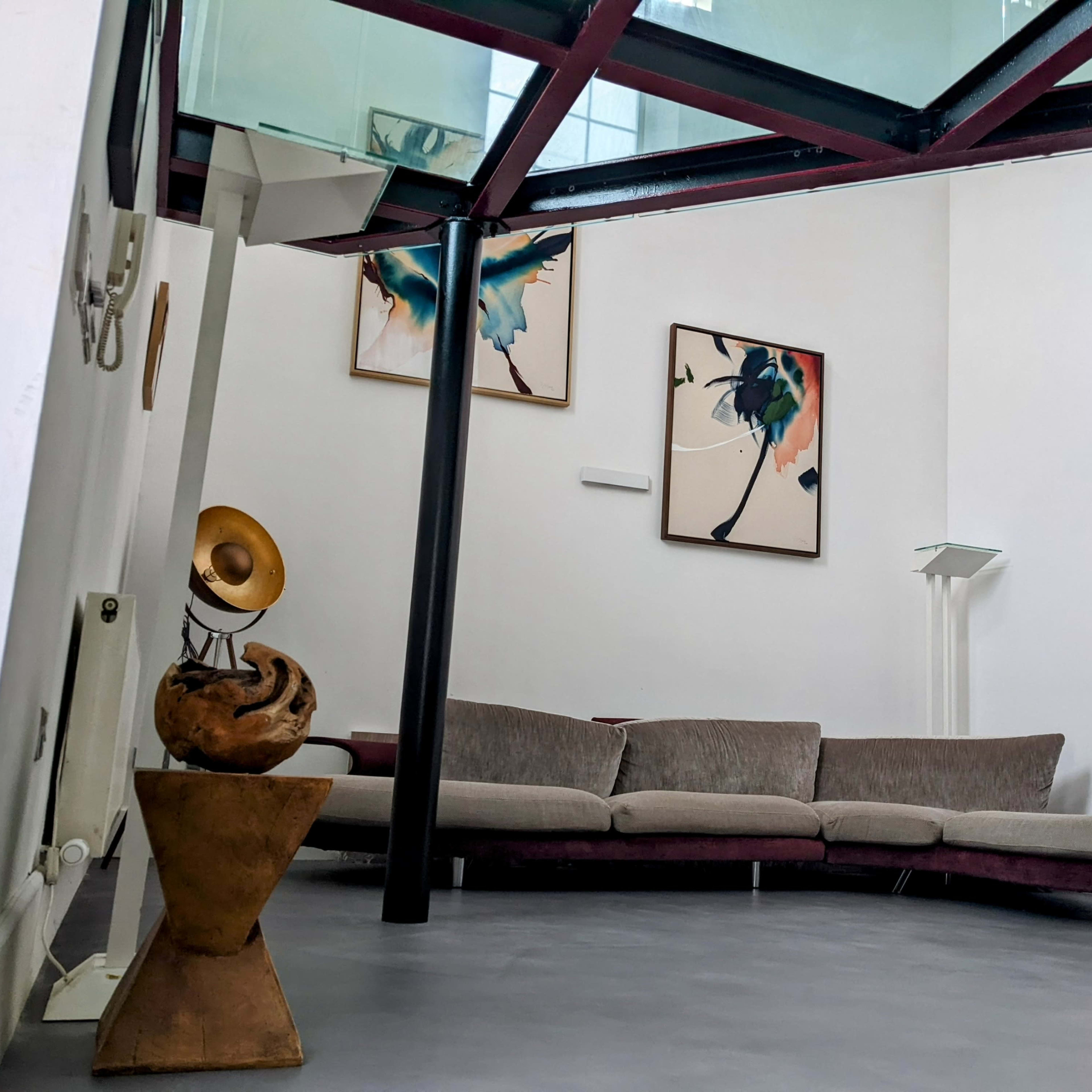 A modern interior with a curved gray sofa, two abstract paintings on the wall, and a glass floor above supported by black pillars.