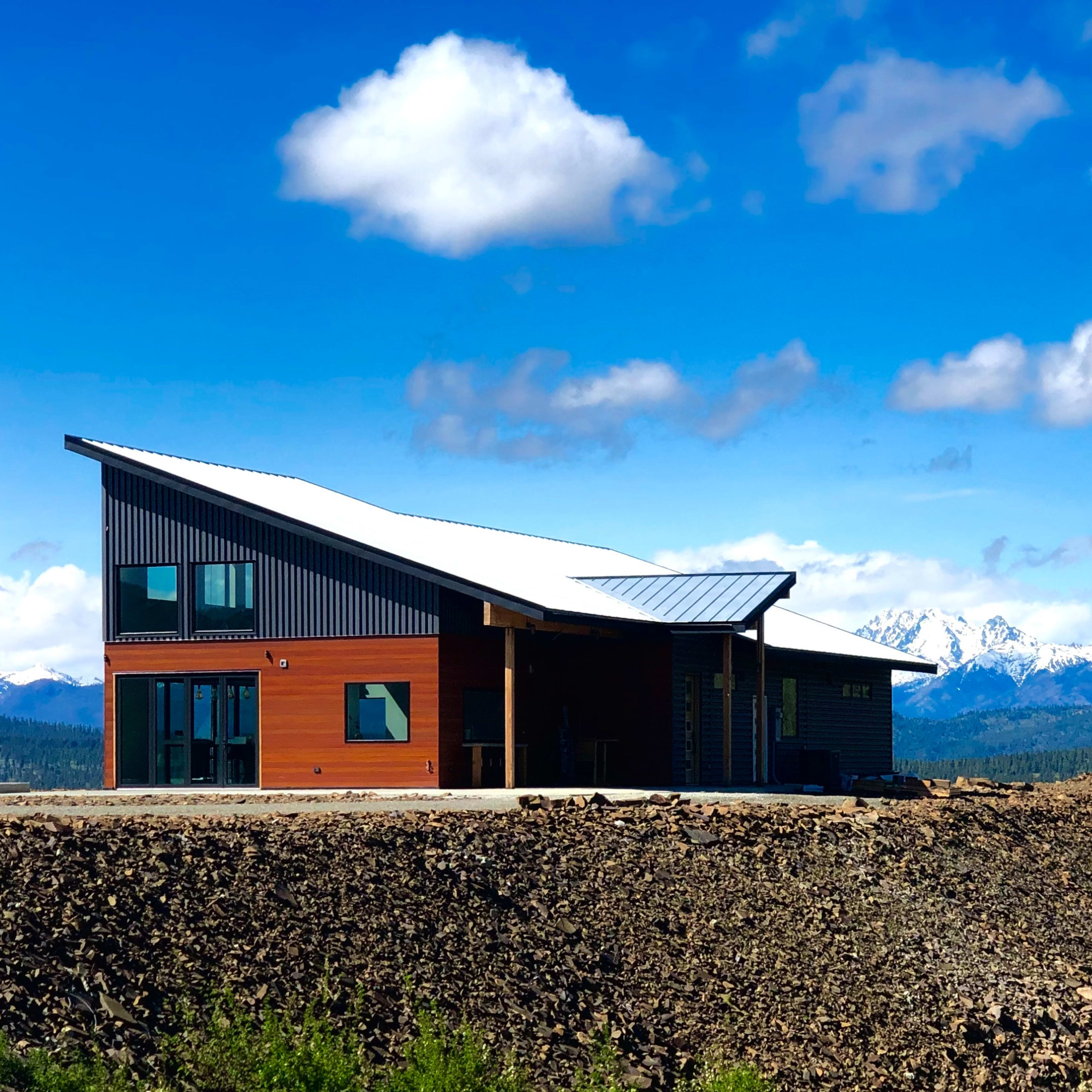 A modern house with a sloped roof situated on a rocky hillside, surrounded by snow-capped mountains under a blue sky with scattered clouds.