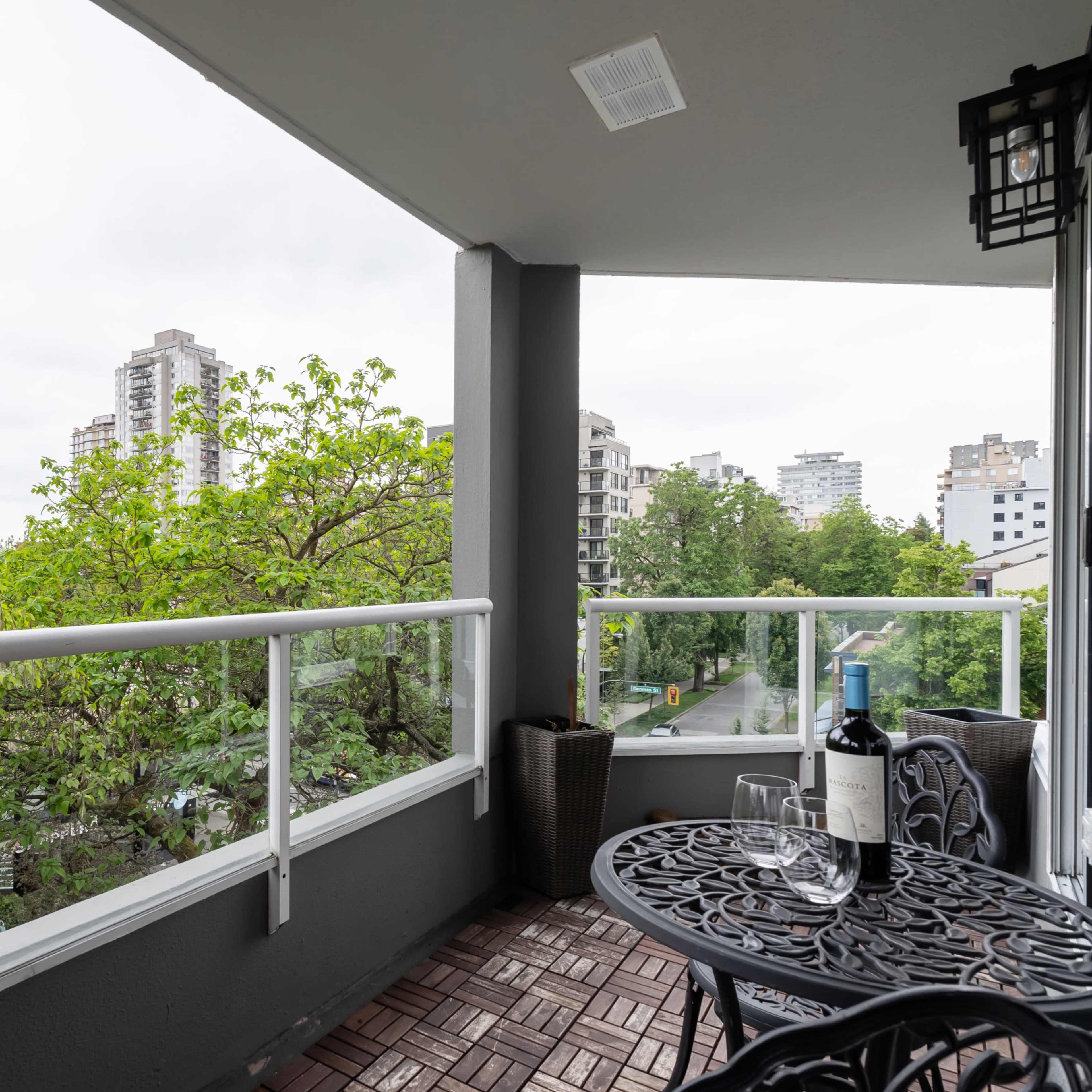 A balcony with a metal table and chairs overlooks a cityscape surrounded by green trees.