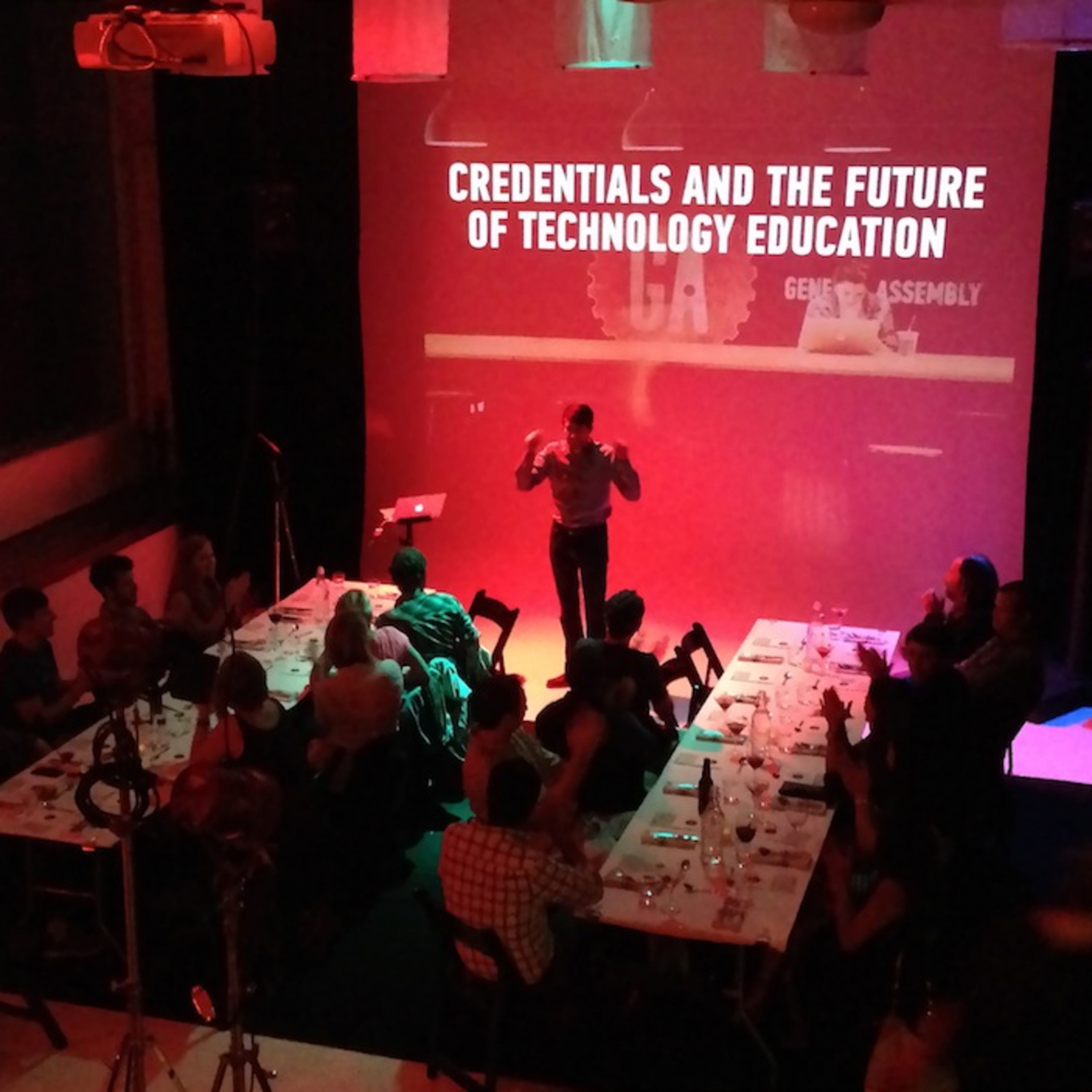 A speaker stands in front of an audience at a dinner event discussing "Credentials and the Future of Technology Education."