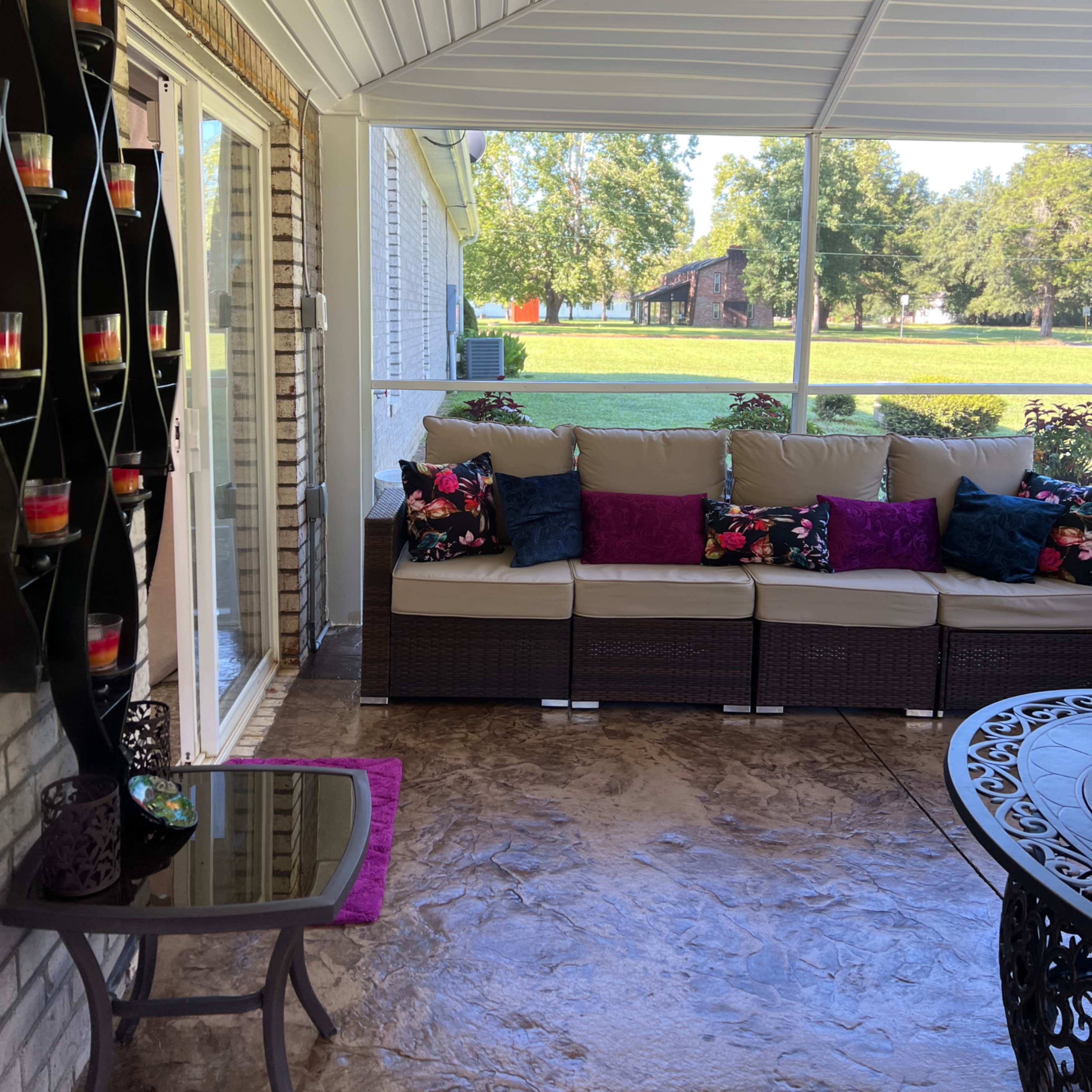 A covered porch with a sofa, decorative pillows, and a table, overlooking a grassy area and trees.