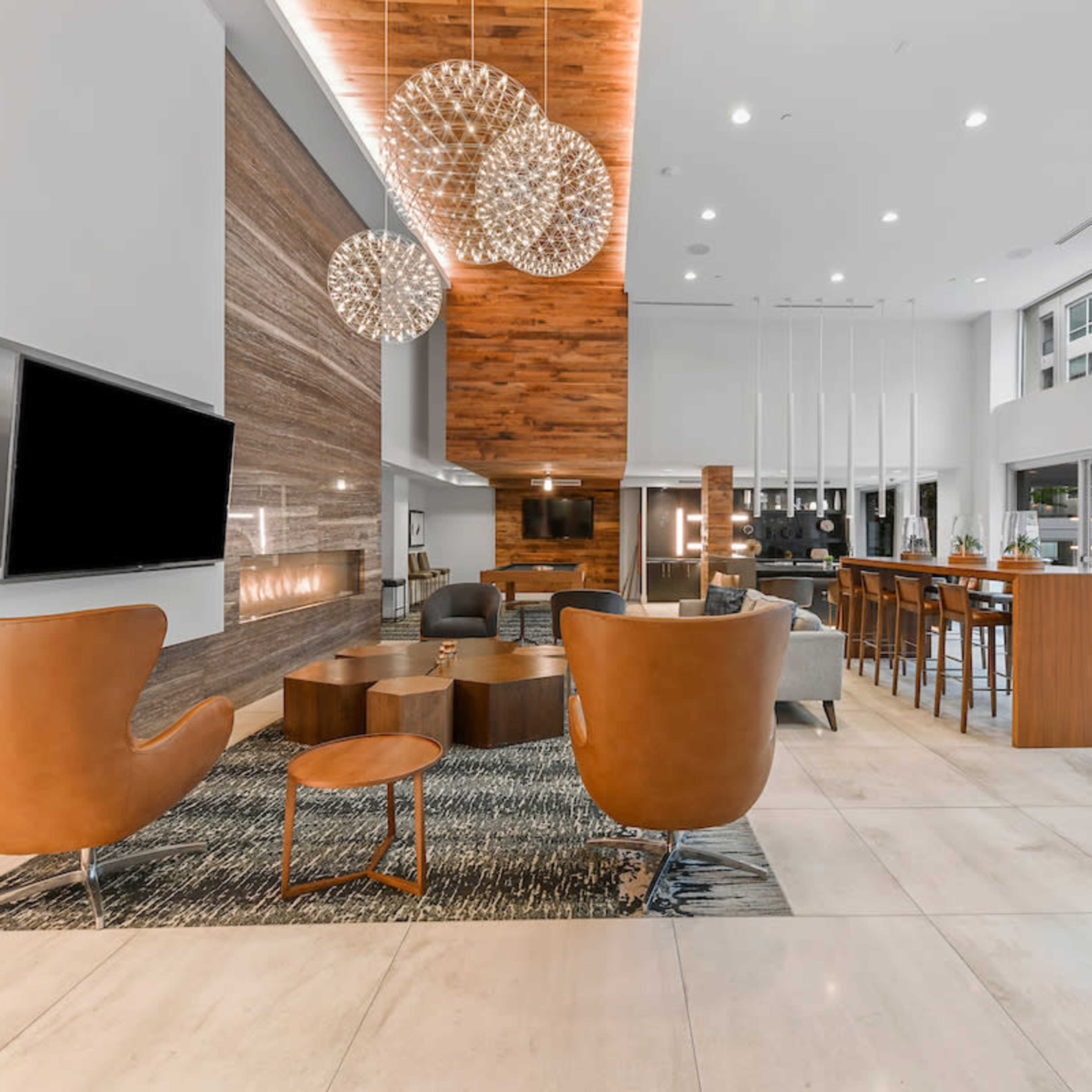A modern lobby with furniture arranged around a central seating area, featuring a television, decorative lighting, and large windows.