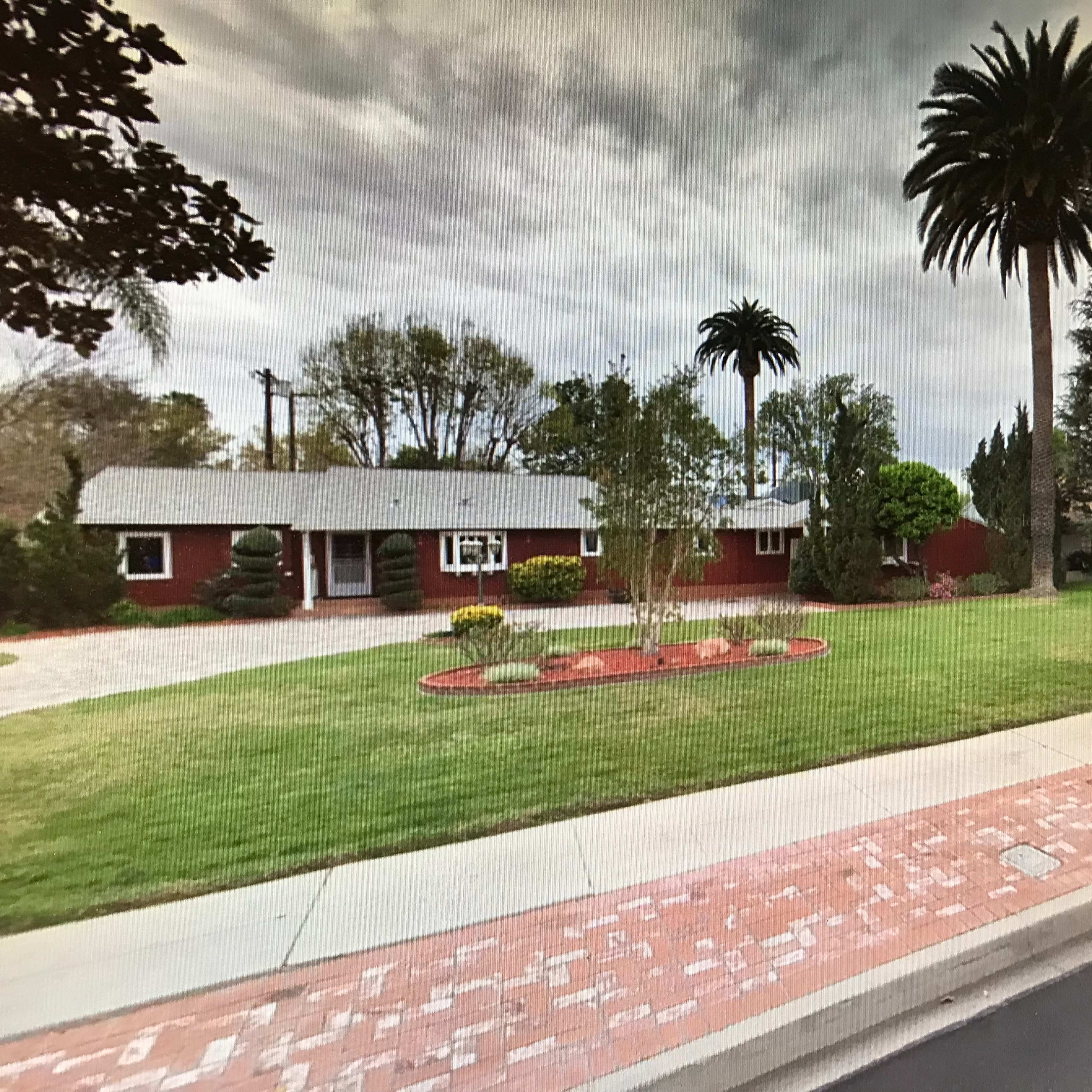A single-story red house with a gray roof is surrounded by a well-manicured lawn and various trees, located on a quiet street.