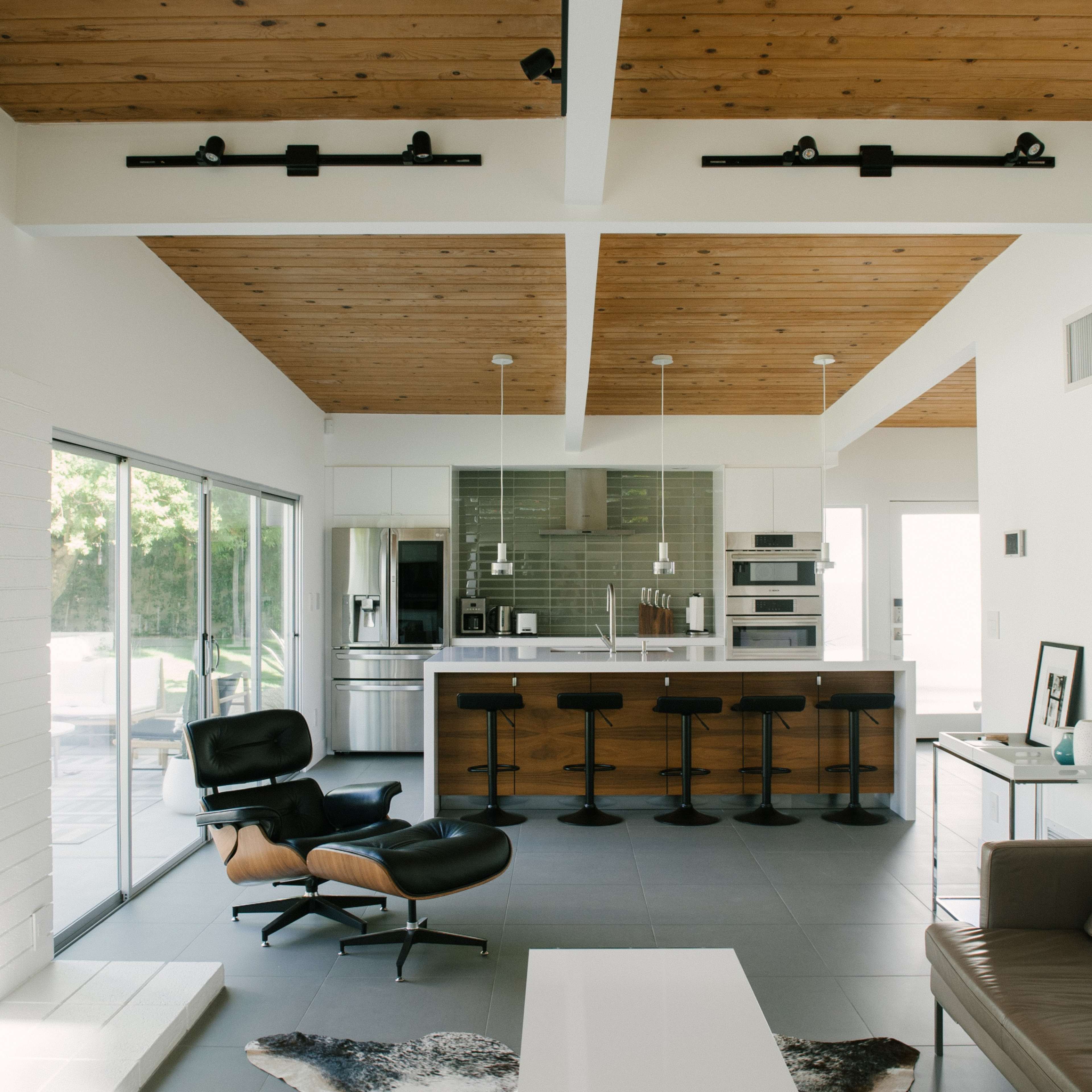 A modern living space with a wooden ceiling, a sleek kitchen in the background, and a leather sofa paired with an iconic chair.