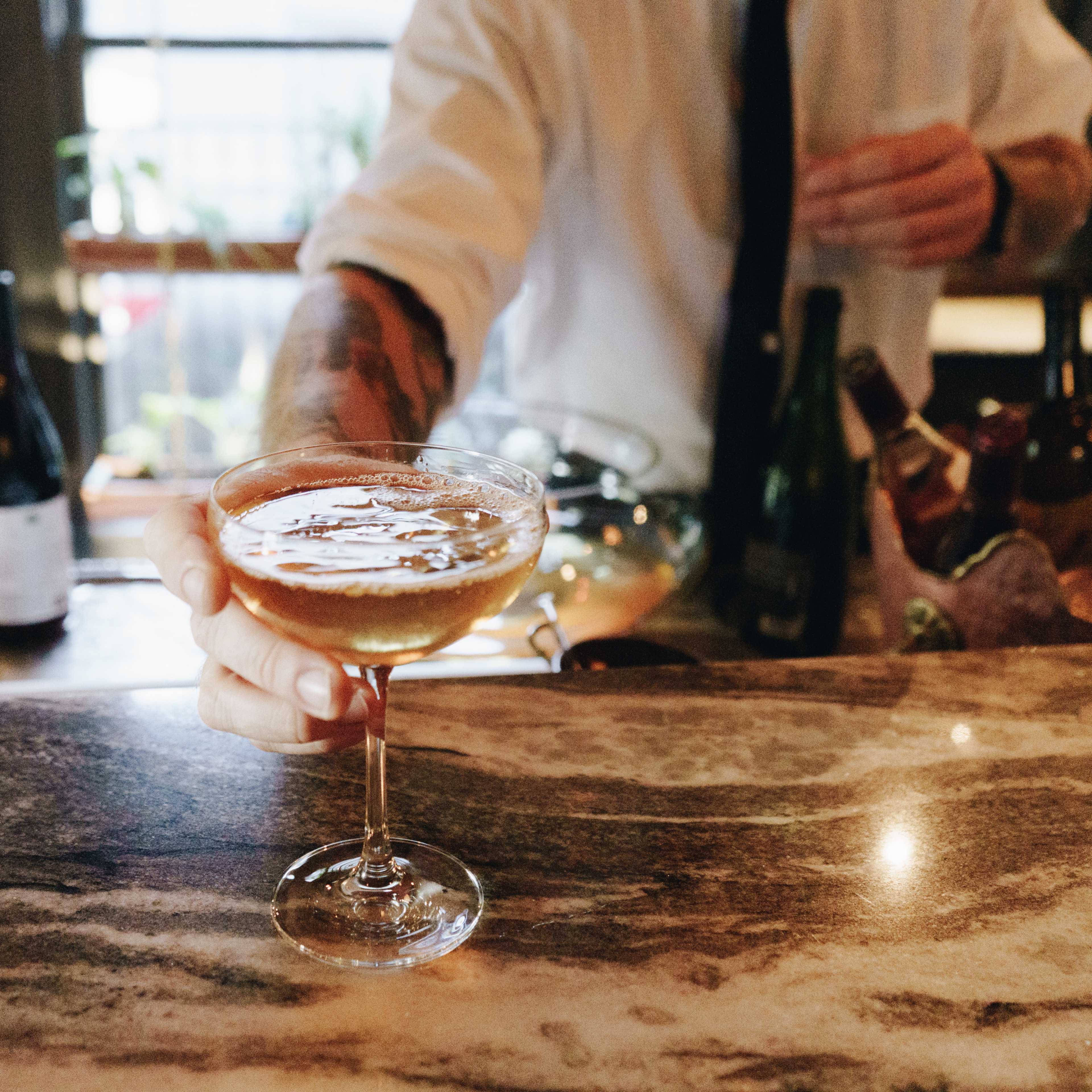 A bartender is serving a cocktail in a coupe glass from behind a marble bar.