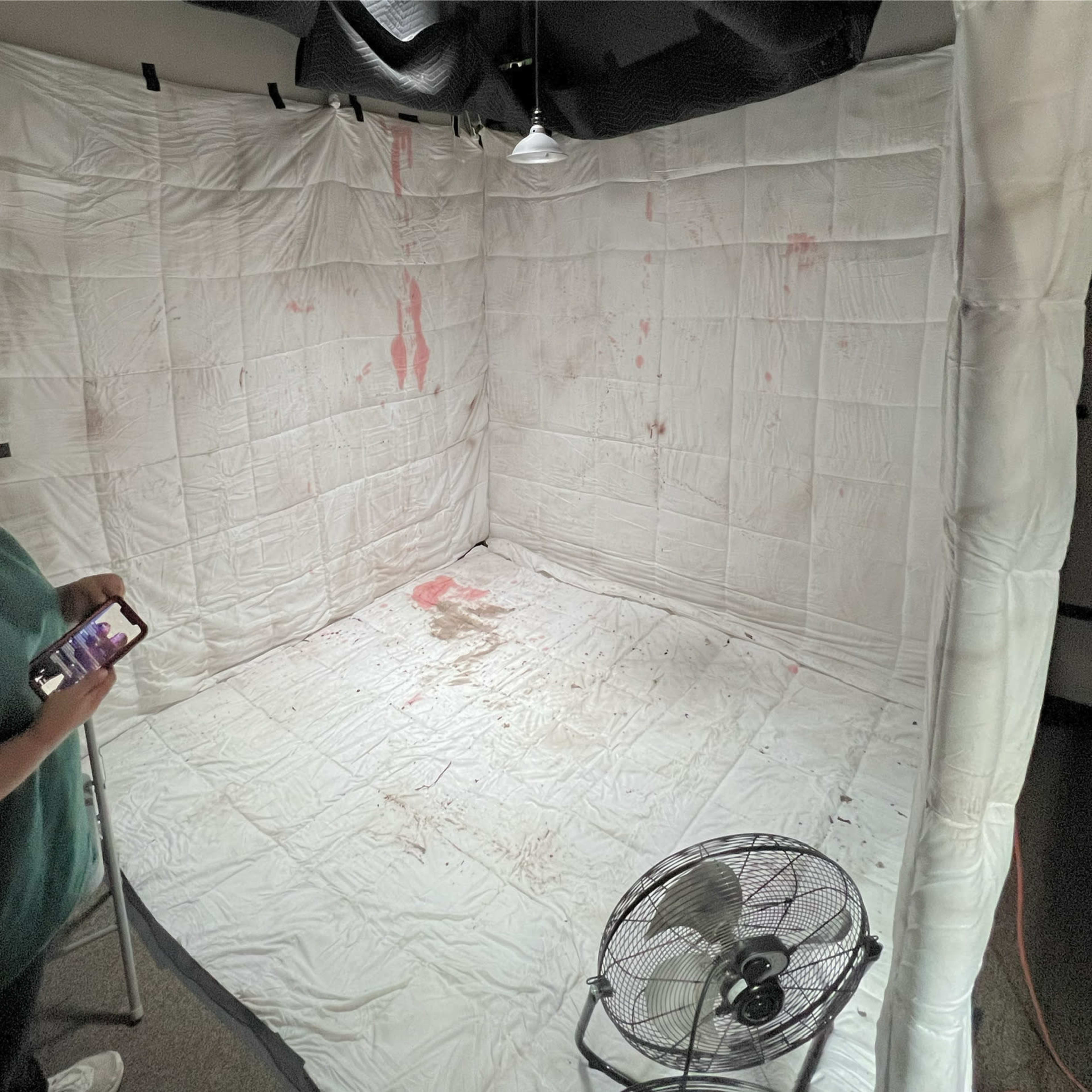 An enclosed room is covered with white blankets, featuring stains on the walls and floor, and a fan in the corner.