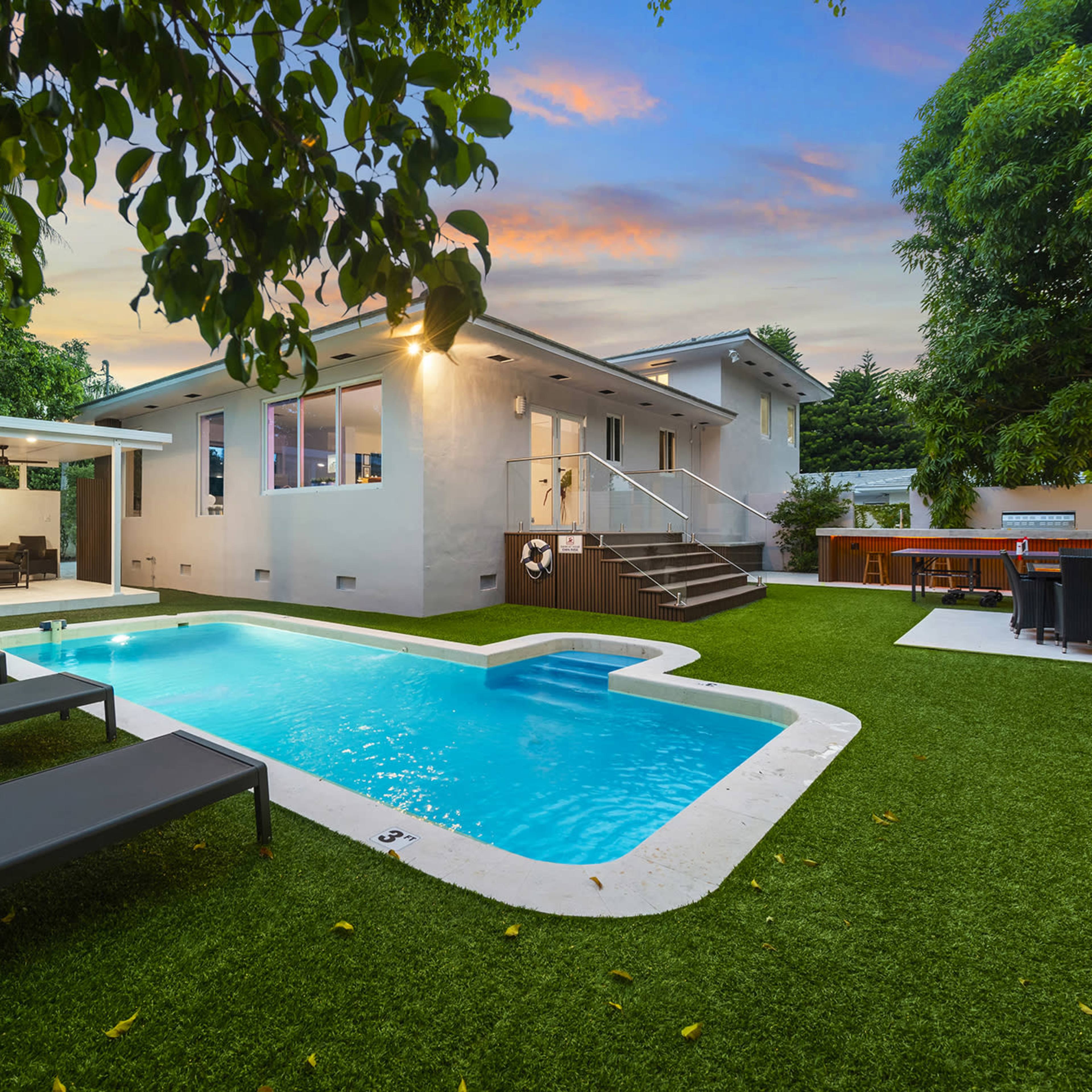 A modern backyard features a swimming pool surrounded by artificial grass, with lounge chairs and a shaded seating area.