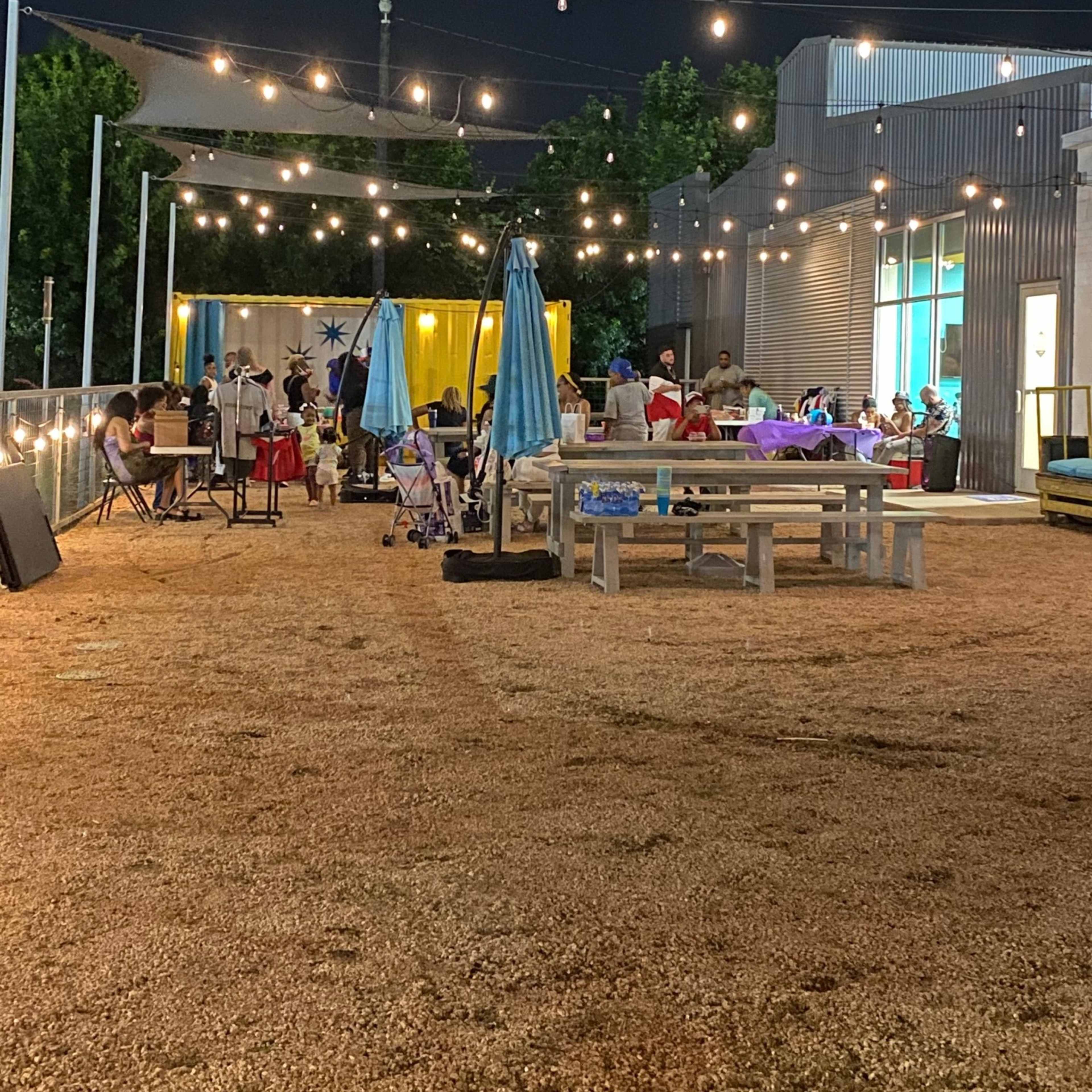 The outdoor area features several tables and chairs under string lights with people gathered in a lively setting at night.