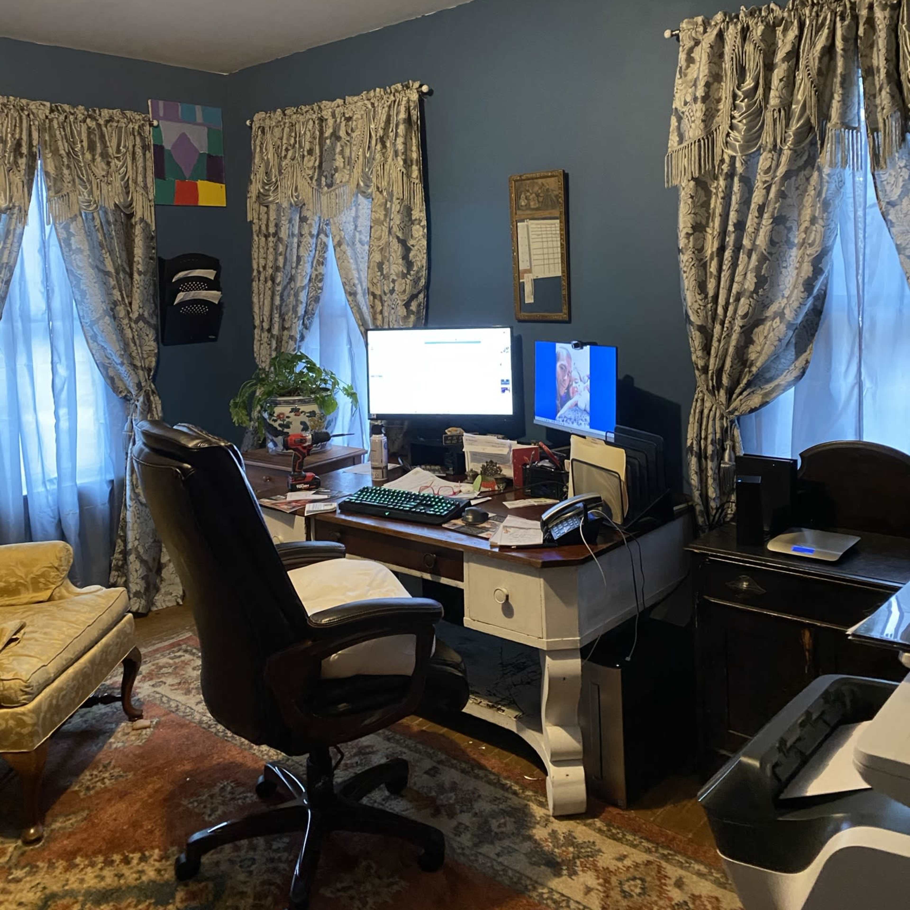 A home office features a desk with a computer, a chair, and large windows dressed with patterned curtains.