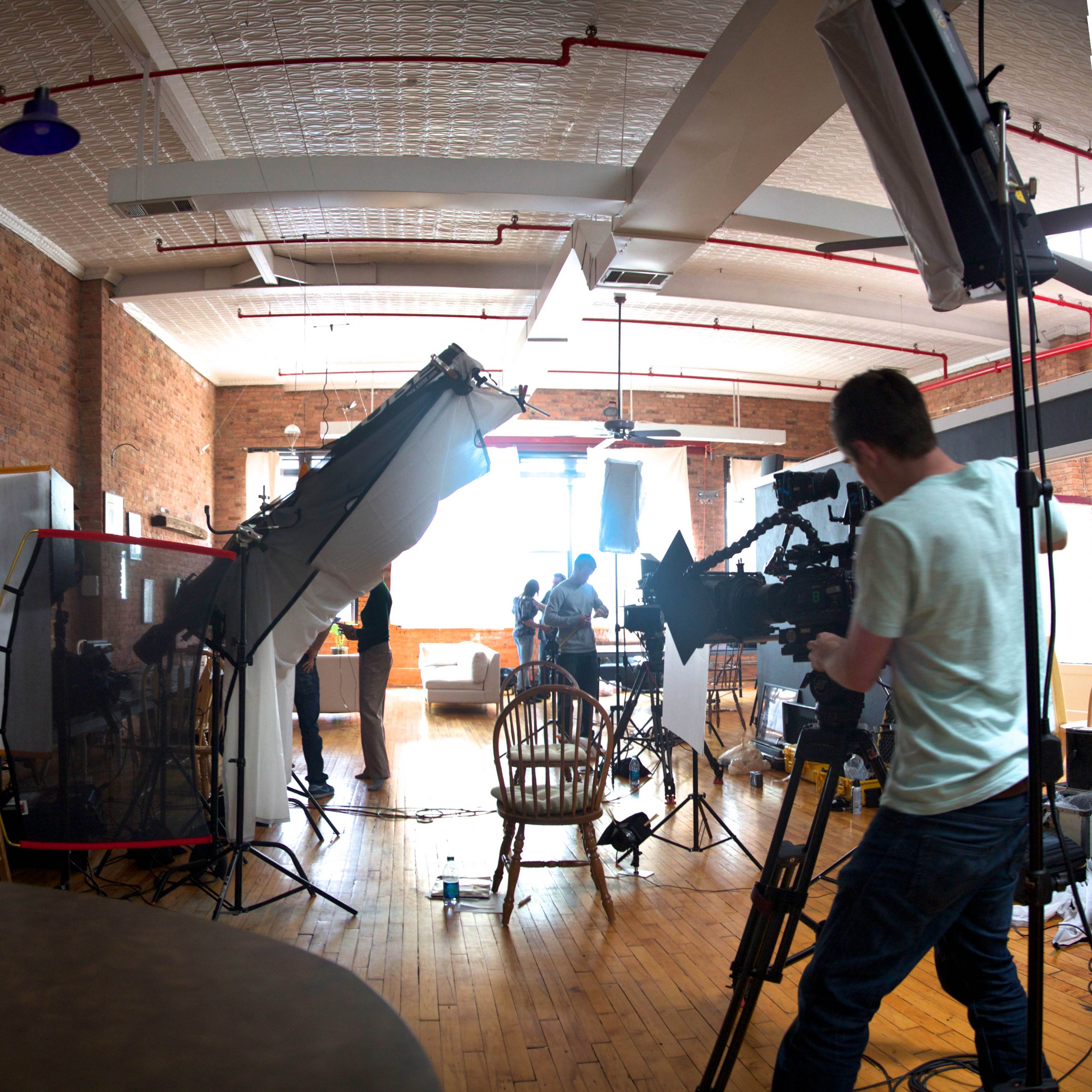A film crew sets up equipment in a spacious, industrial-style studio with exposed brick walls and large windows.