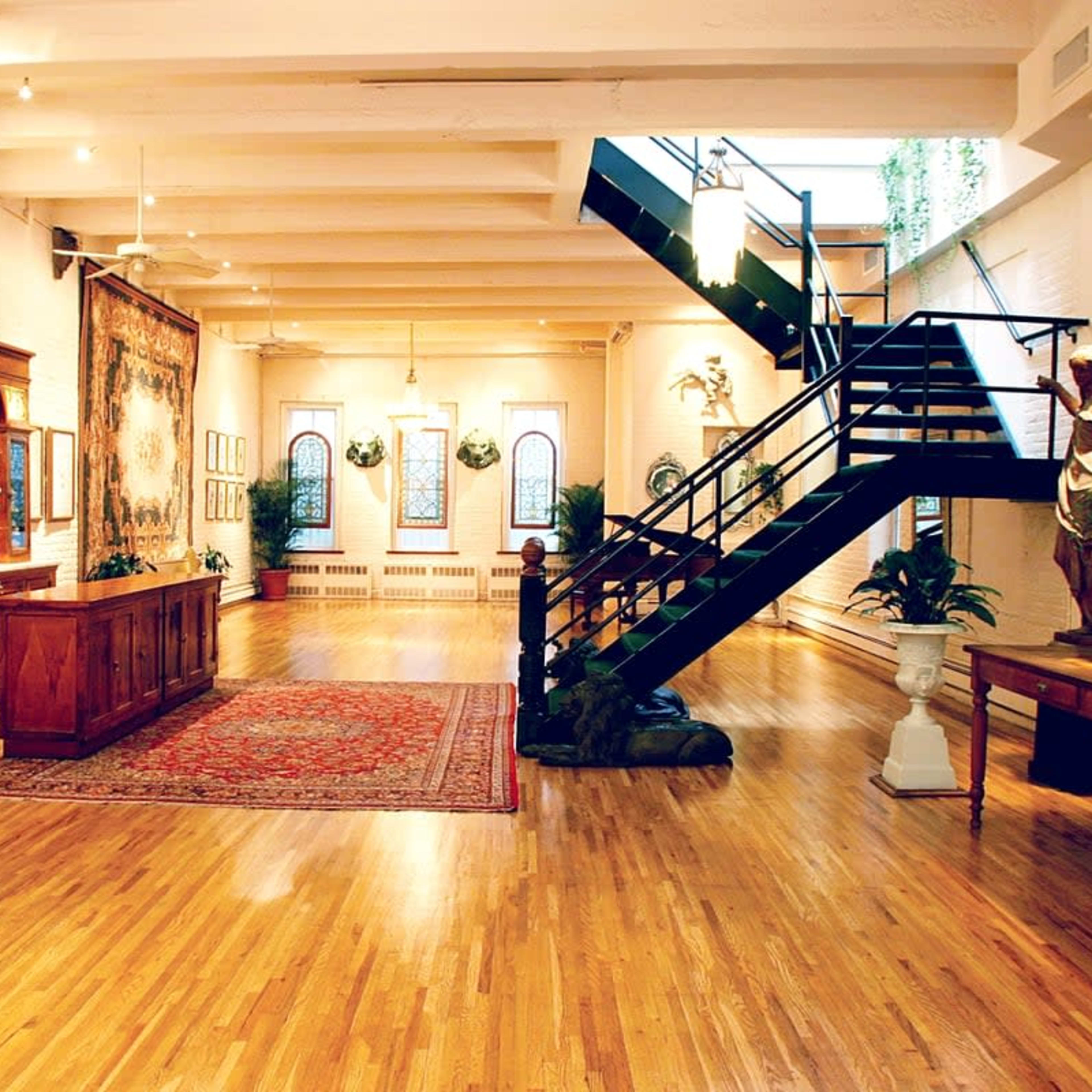 A spacious art gallery with wooden floors, a decorative rug, large windows, and a staircase leading to an upper level.