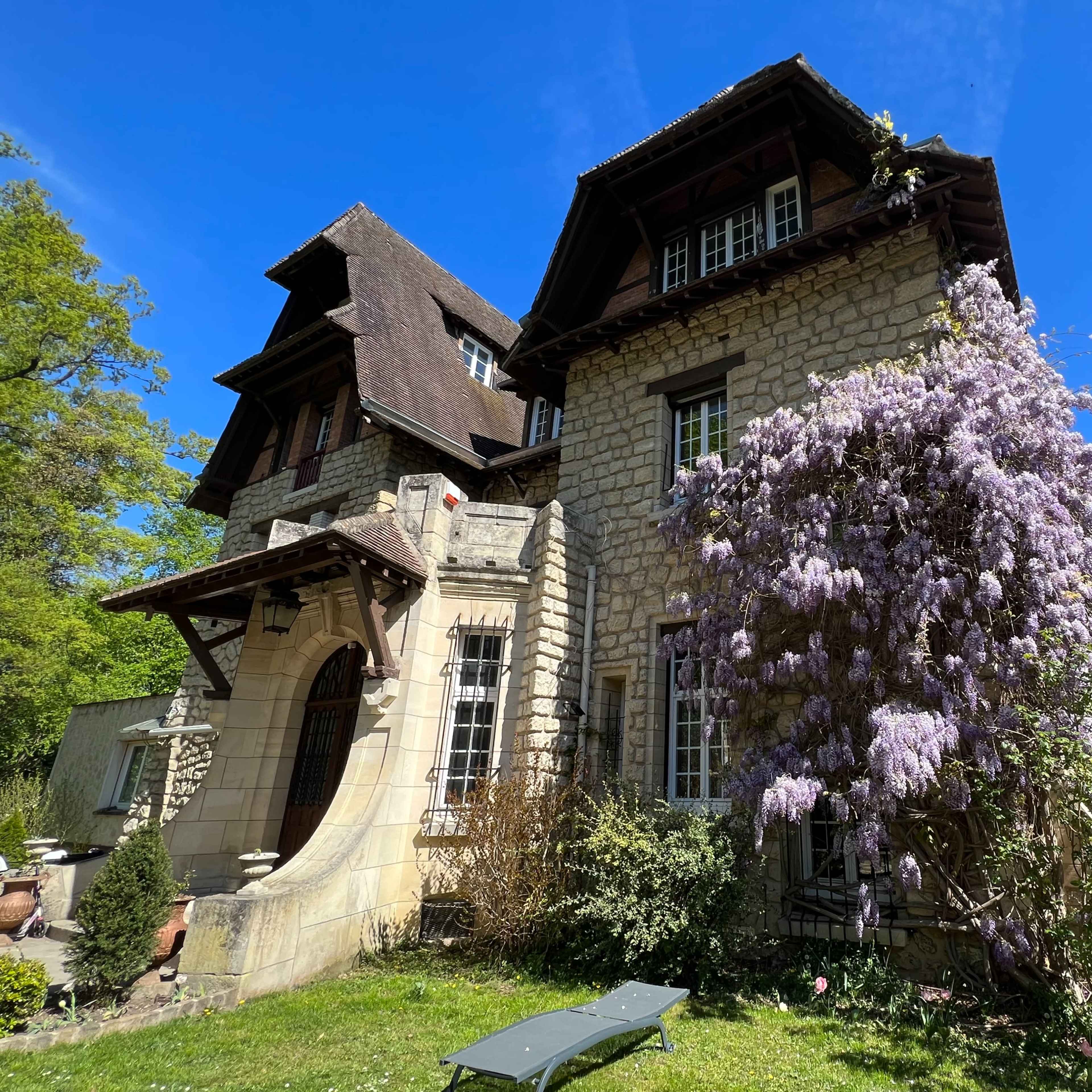 A stone house with a sloping roof is adorned with blooming wisteria and surrounded by green foliage under a clear blue sky.