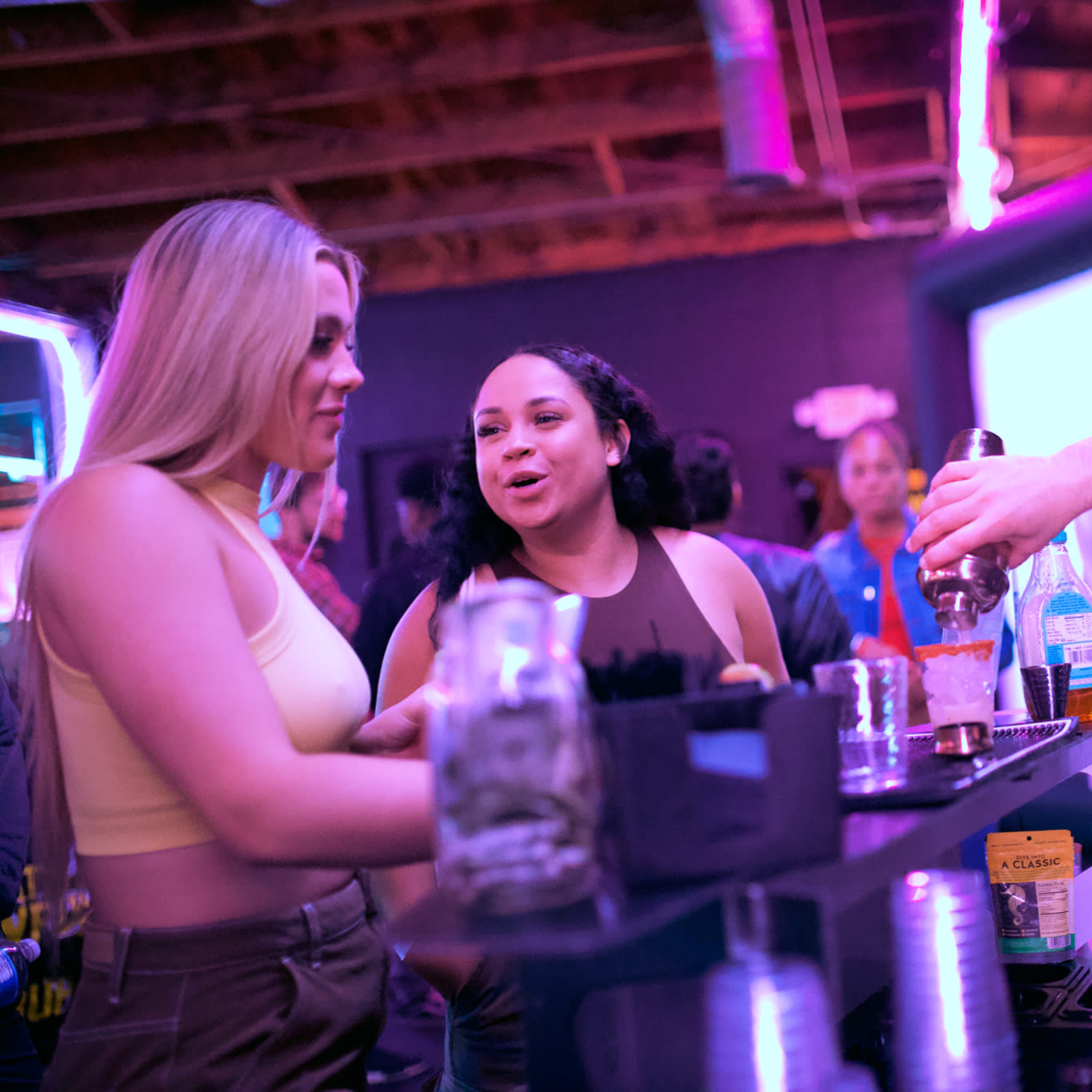 A bartender prepares drinks while two women engage in conversation at a lively bar.